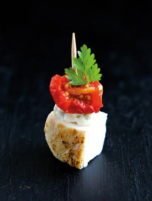 En elegant bid af en lille appetitvækker med grillet kylling, toppet med en skive tørret tomat, en creme og et stykke persille, stukket på en tandstik.