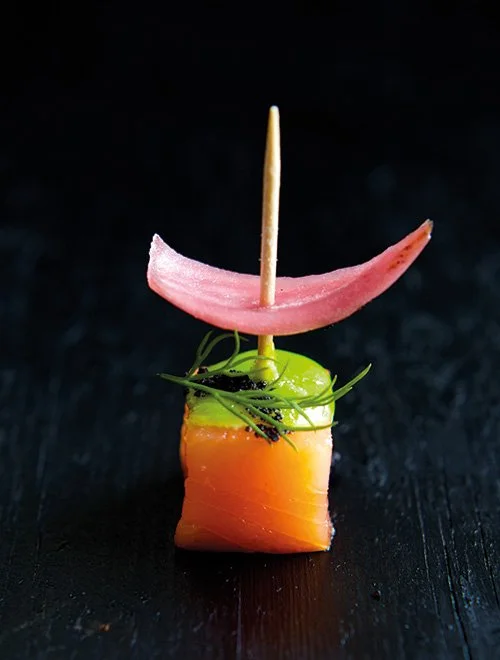En elegant madbid med laks, grøn sauce, urter og en pink skive ovenpå, samlet med et spyd.