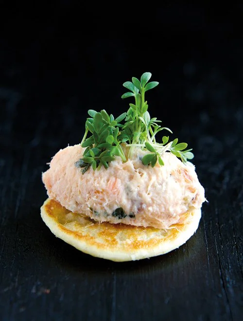 En blinis toppet med laksemousse og frisk karse på en mørk baggrund.
