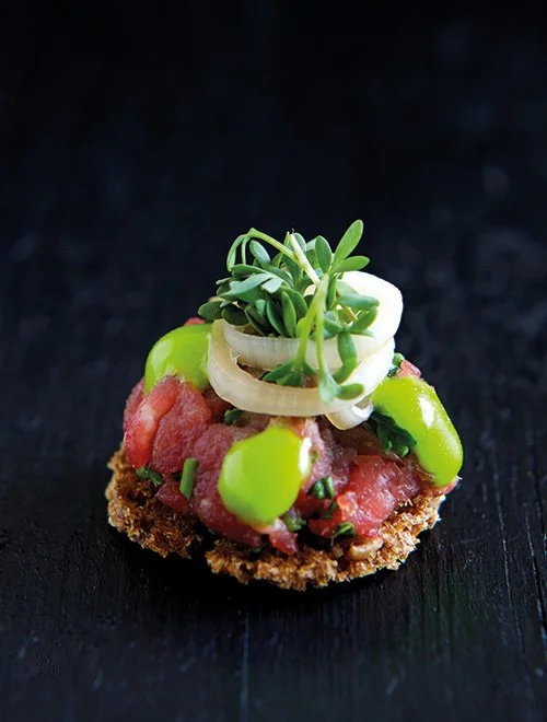 Lille smørrebrød med råt kød, grønne urter, løg og grønne sauceklatter på en mørk overflade.