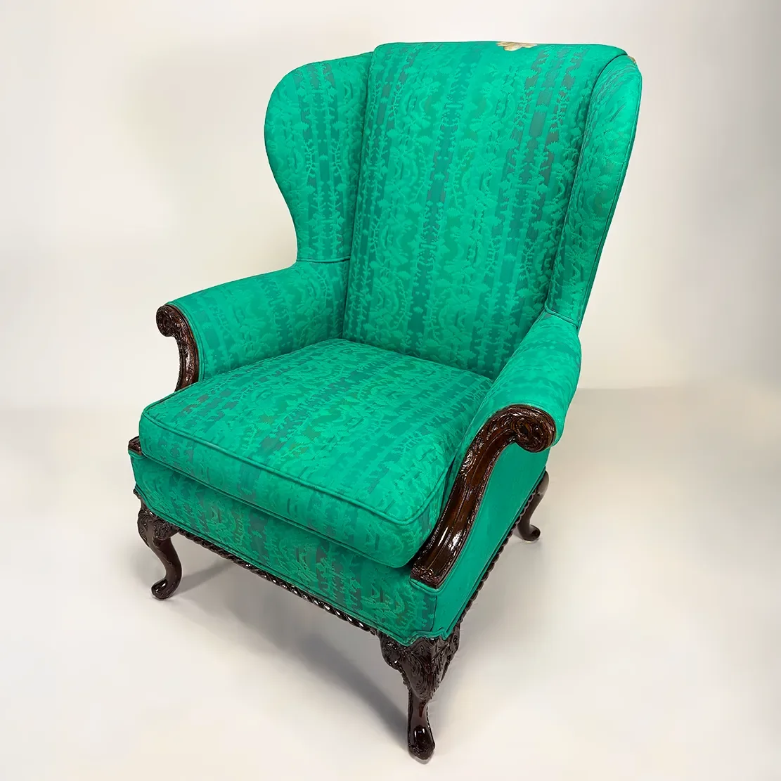 An antique wingback armchair with green patterned upholstery and dark wooden carved legs and armrests.