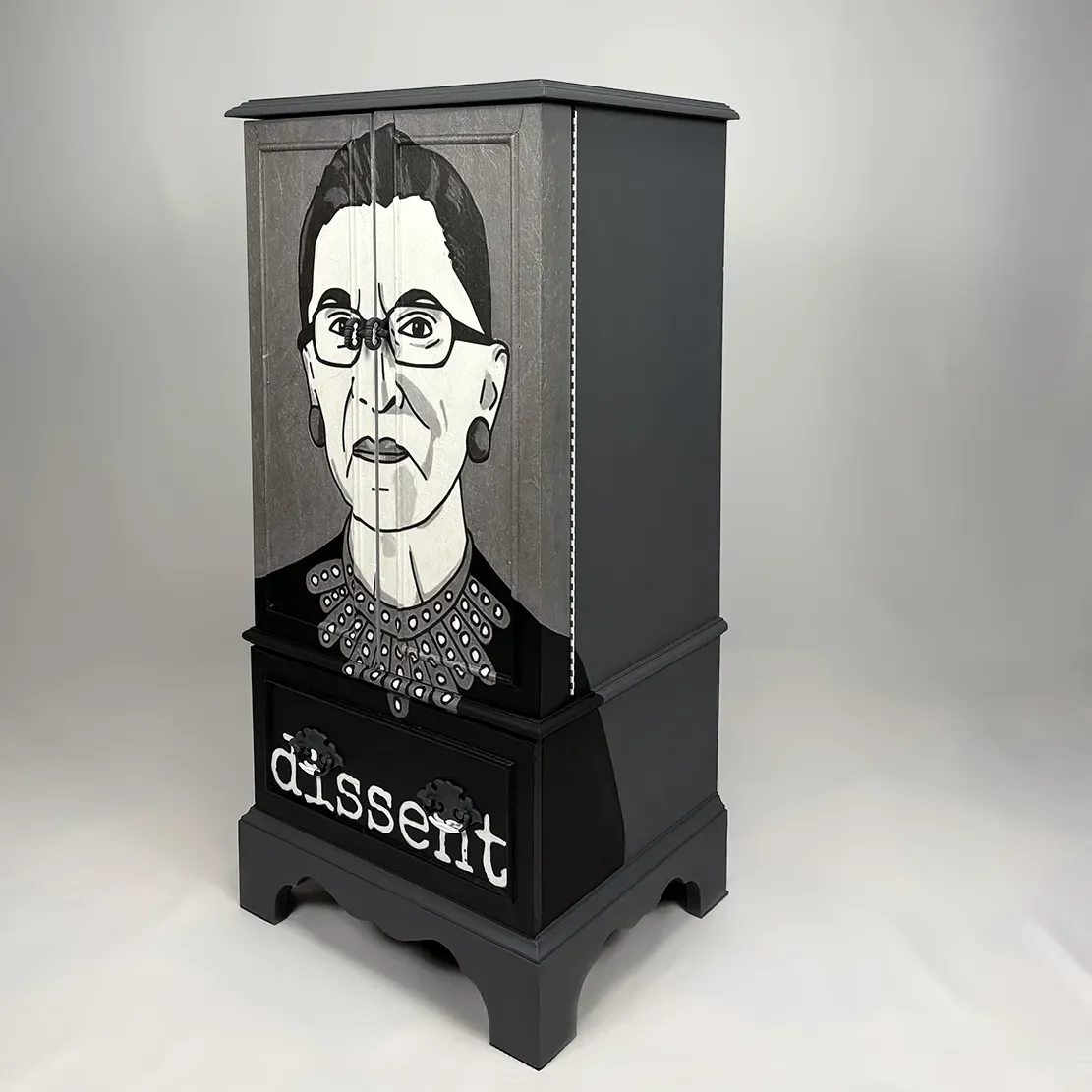 A black cabinet with a painted portrait of Ruth Bader Ginsburg, featuring her glasses, earrings, and distinctive collar, with the word 'dissent' written at the bottom.