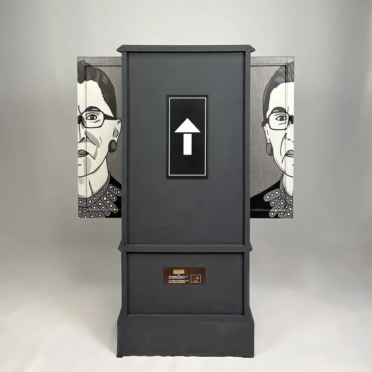 A black elevator with a white upward arrow sign on the door, flanked by black and white abstract portraits of women wearing glasses and earrings.