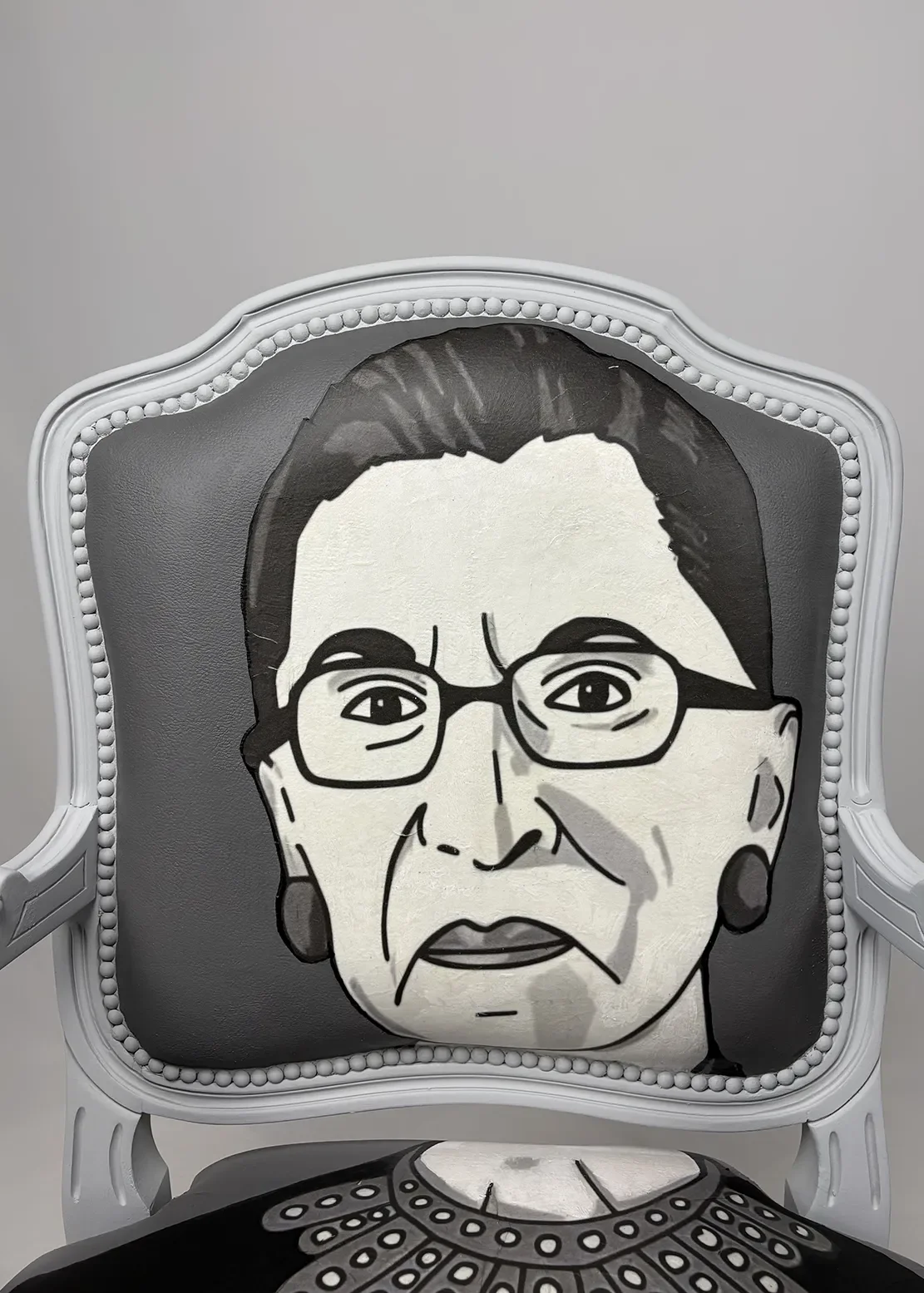 A chair with a cushion featuring a painted portrait of an elderly woman with glasses, earrings, and a solemn expression, set against a plain wall.