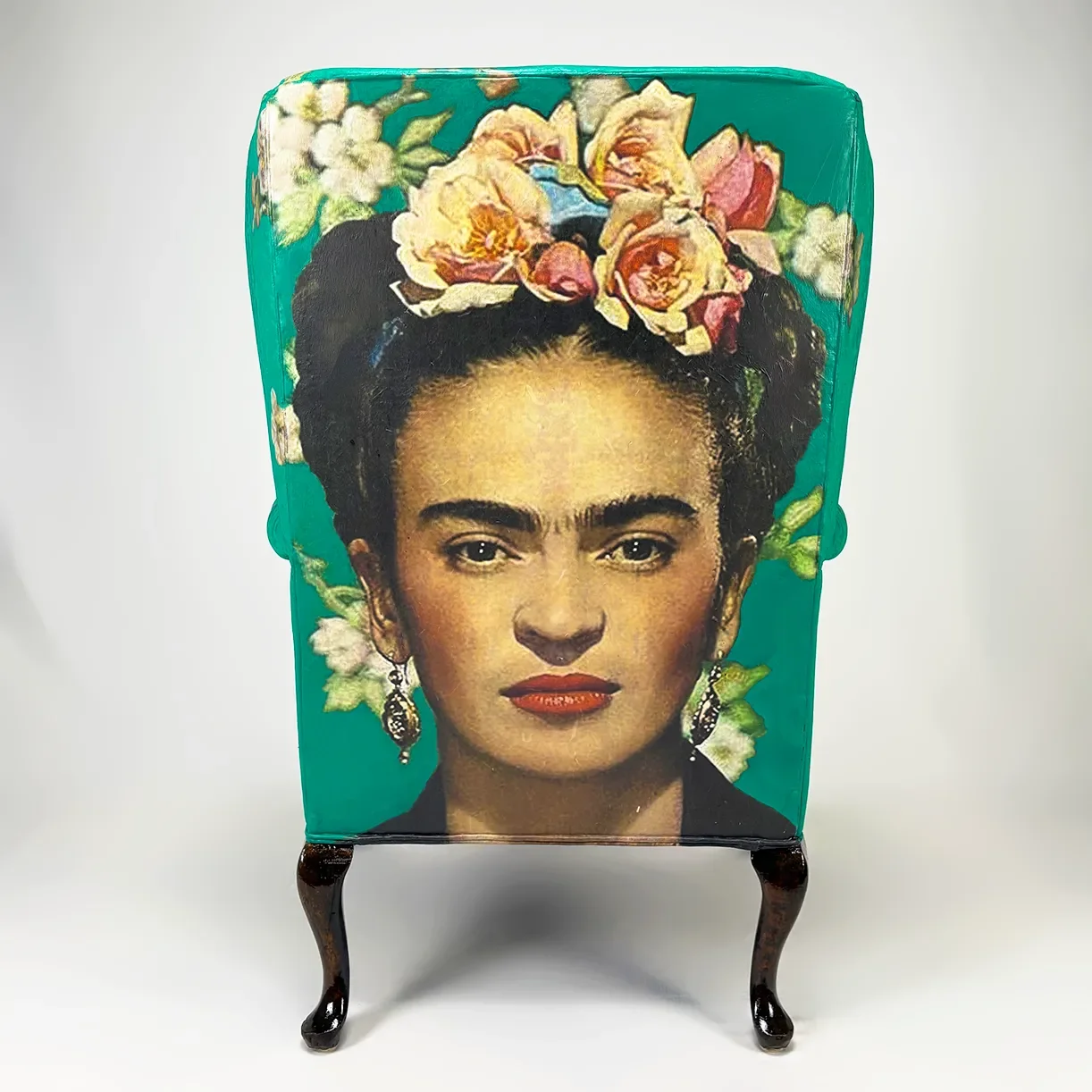 A back of a redesigned vintage wingback chair with decoupage artwork of Frida Kahlo with flowers, wearing earrings, and bright red lipstick, against a teal background