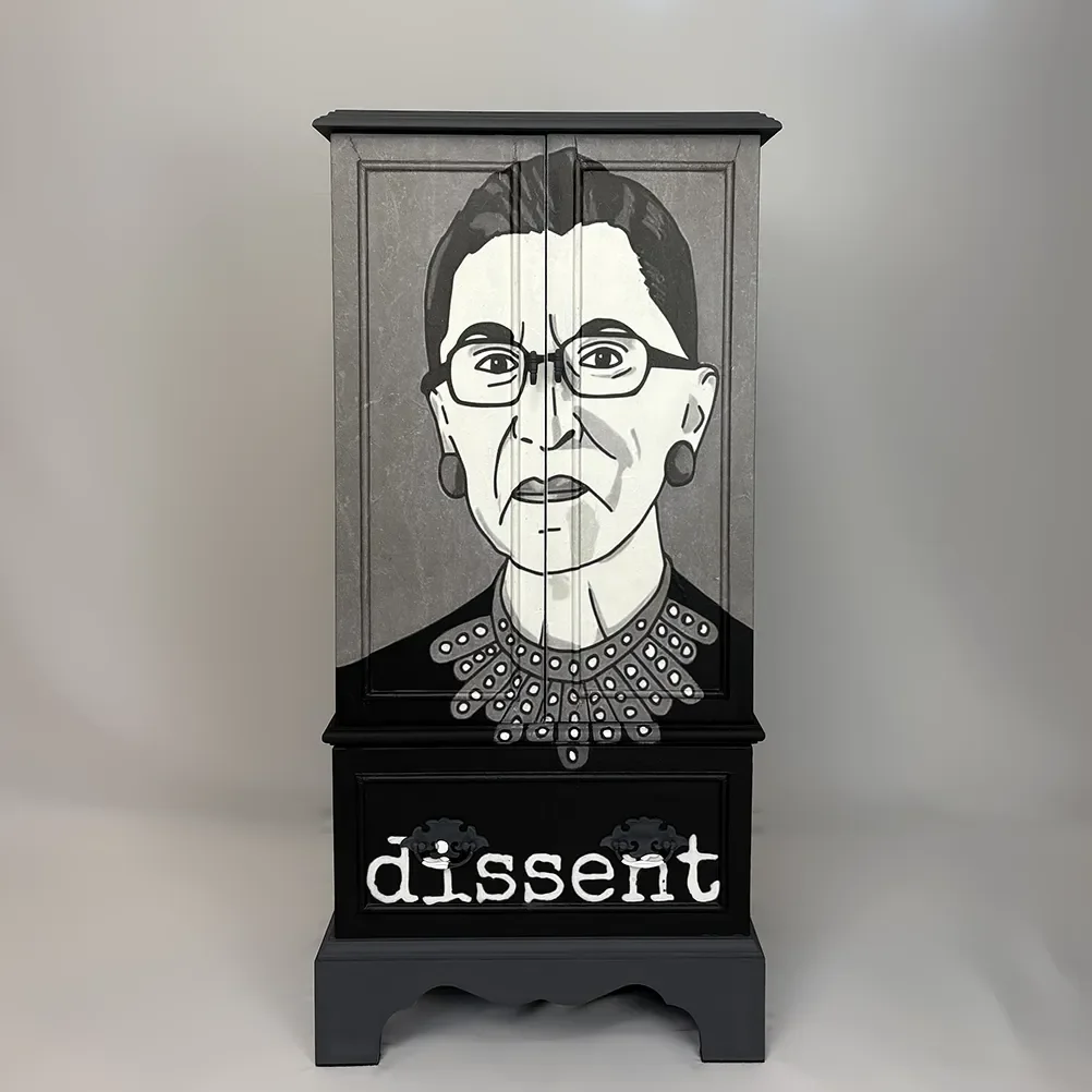 A black cabinet with a hand-painted portrait of Ruth Bader Ginsburg, including her glasses, earrings, and iconic collar. The word "dissent" is painted below her portrait.