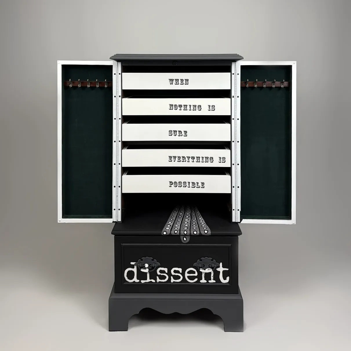 A black and white storage cabinet with open doors, displaying drawers with words: 'When nothing is sure everything is possible.' The word 'dissent' is written on the lower part of the cabinet.