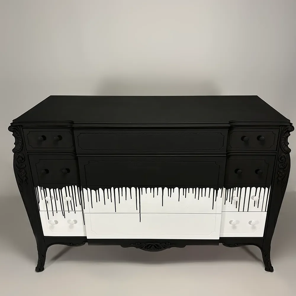 A black dresser with ornate carved details on the sides and legs, featuring a white drawer in the center with a black dripping paint design.
