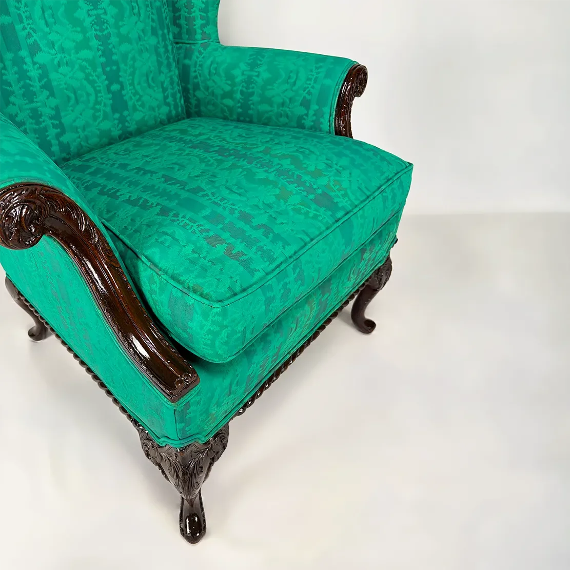 Close-up of a vintage green upholstered armchair with dark wooden carved accents and curved legs, set against a plain white background.