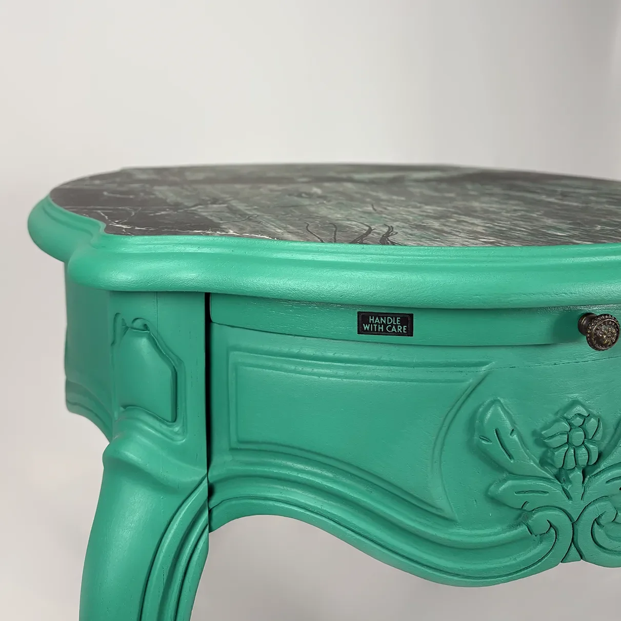 A turquoise vintage nightstand with decorative carvings, a black handle with a label saying 'HANDLE WITH CARE', and a marble top.