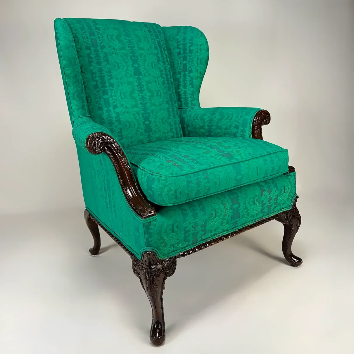 Green upholstered armchair with dark wooden carved legs and arm accents, against a plain light background.