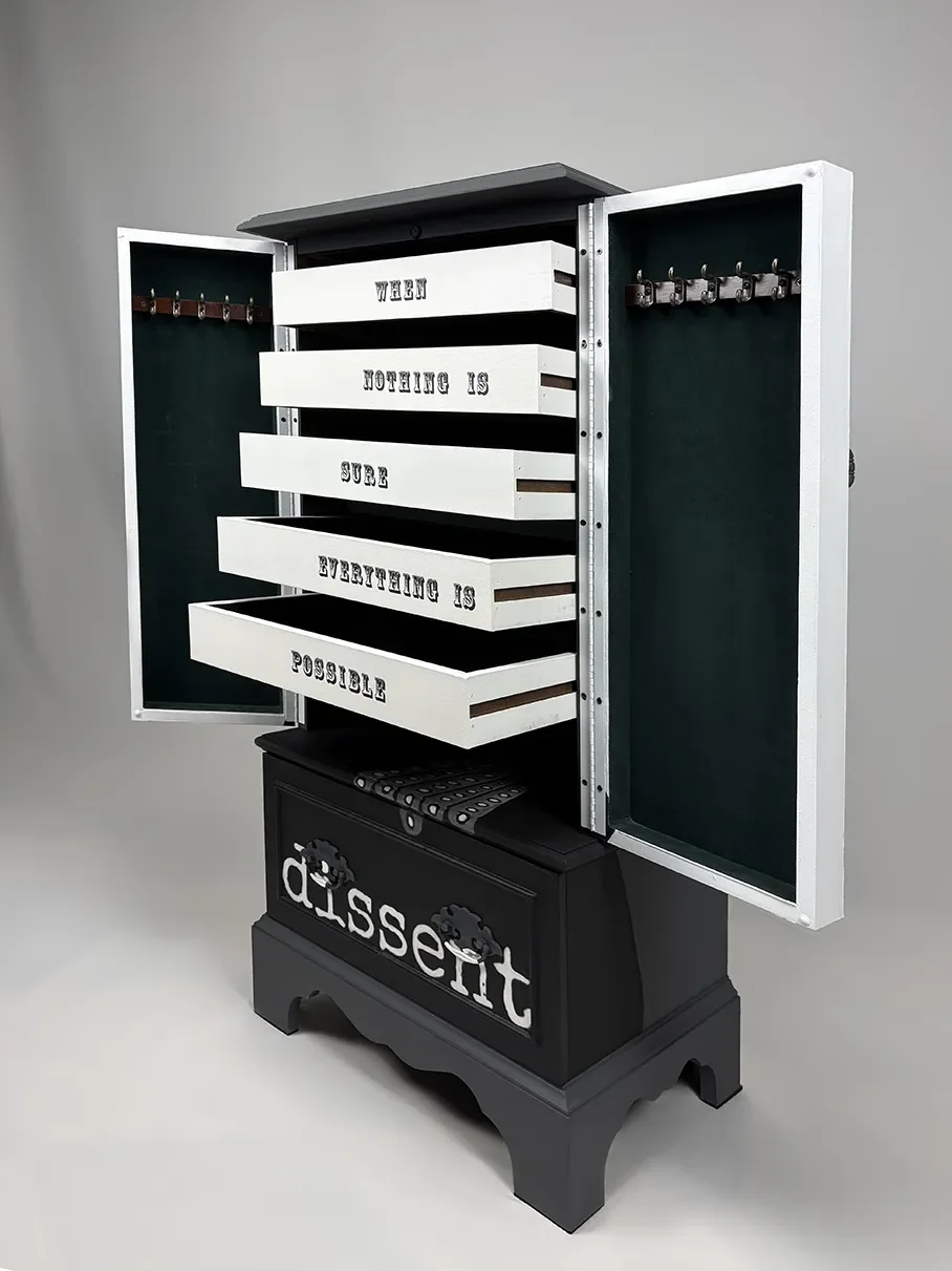 A decorative cabinet with open doors revealing pull-out drawers with the quote, 'When nothing is sure, everything is possible.' The cabinet has a black base with the word 'dissent' in white, and the drawers are white with black text.