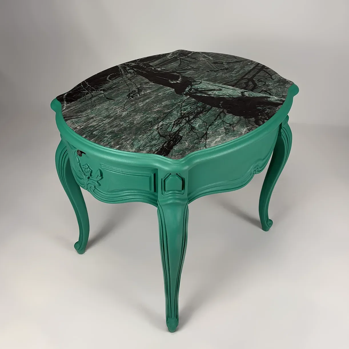A small, ornate table with a dark green marble top featuring black and white veining. The table has a bright turquoise, carved wooden base with curved legs and decorative details.
