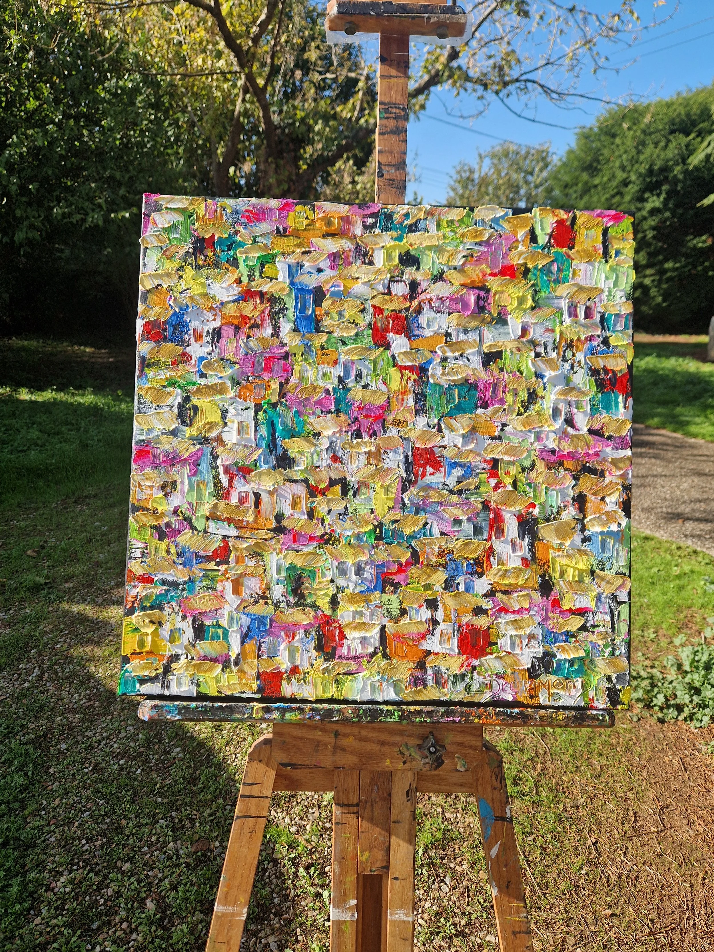 Por Entre as Vielas, colourful Brazilian favela oil painting by Christine Drummond, 60 × 60 cm, created with a palette knife and displayed on an easel outdoors.