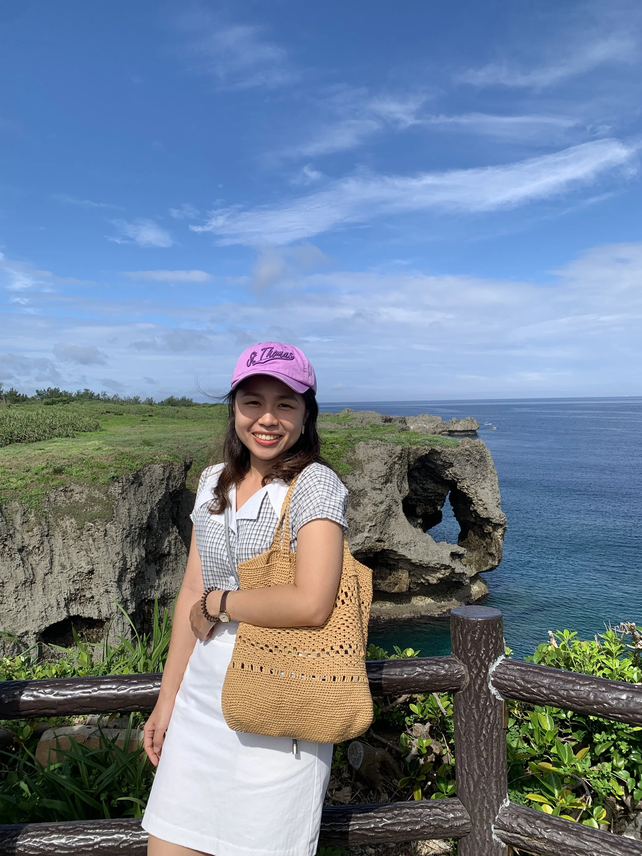 A smiling woman wearing a pink cap, white dress, and a crochet bag standing outdoors near the ocean with rocks and green vegetation, under a partly cloudy blue sky.