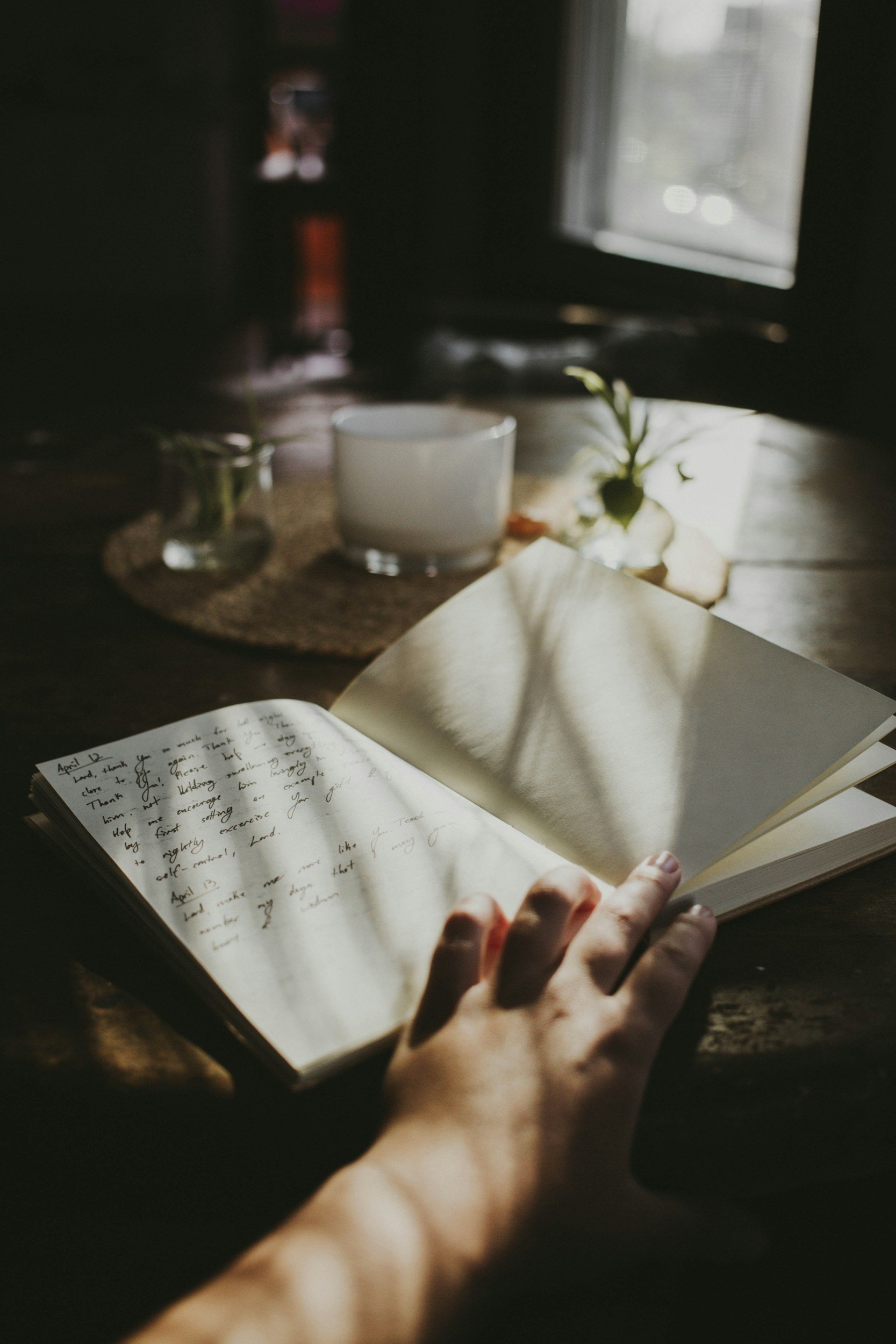 A person holding a journal or notebook with handwritten notes in one hand, while sitting at a dark wooden table. In the background, there are two small plants, a white mug, and a computer monitor near a window with natural light streaming in.