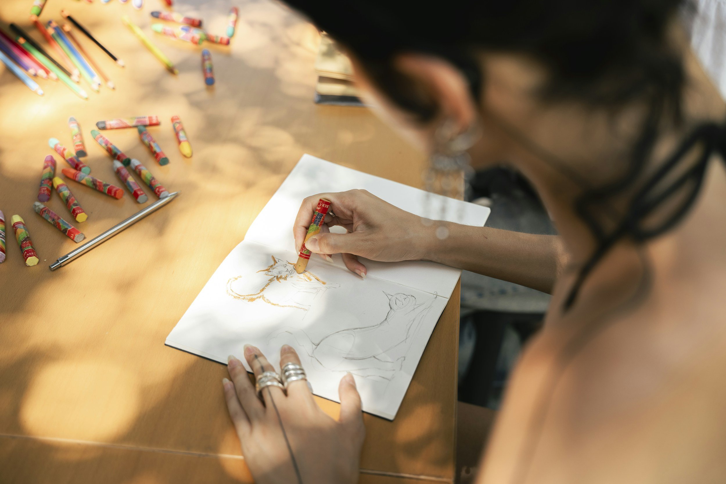A person drawing a horse in a sketchbook on a wooden table, with colorful markers and crayons scattered around.