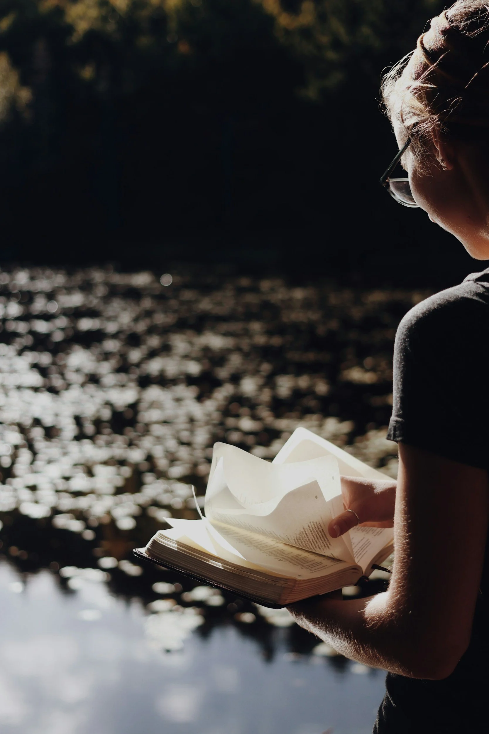 A person with glasses reading a book outdoors near a body of water, with sunlight illuminating their hair and hand holding the open book.
