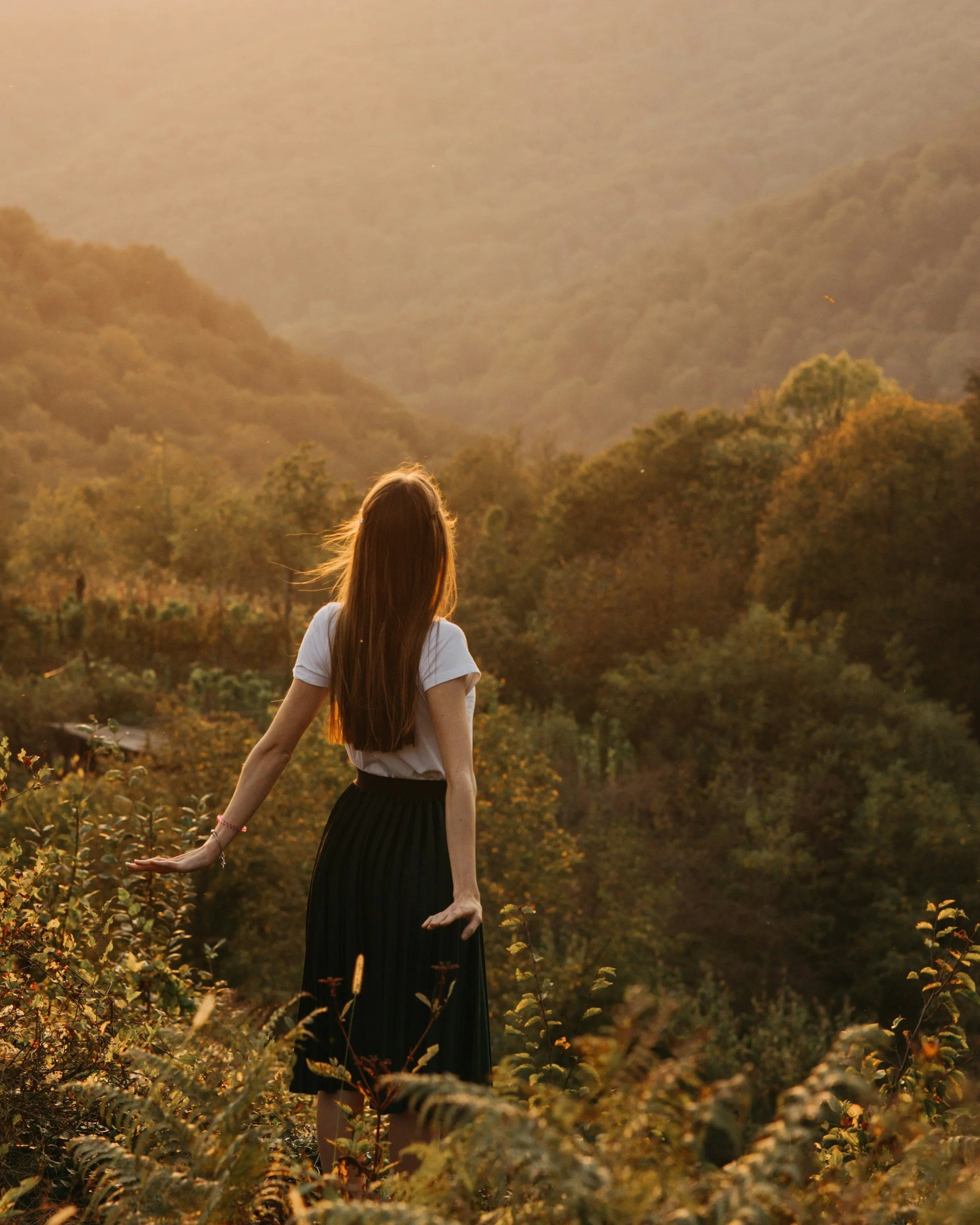 A woman with long brown hair wearing a white shirt and black skirt standing in a field of plants with her arm extended, facing away, during sunset with mountains and trees in the background.