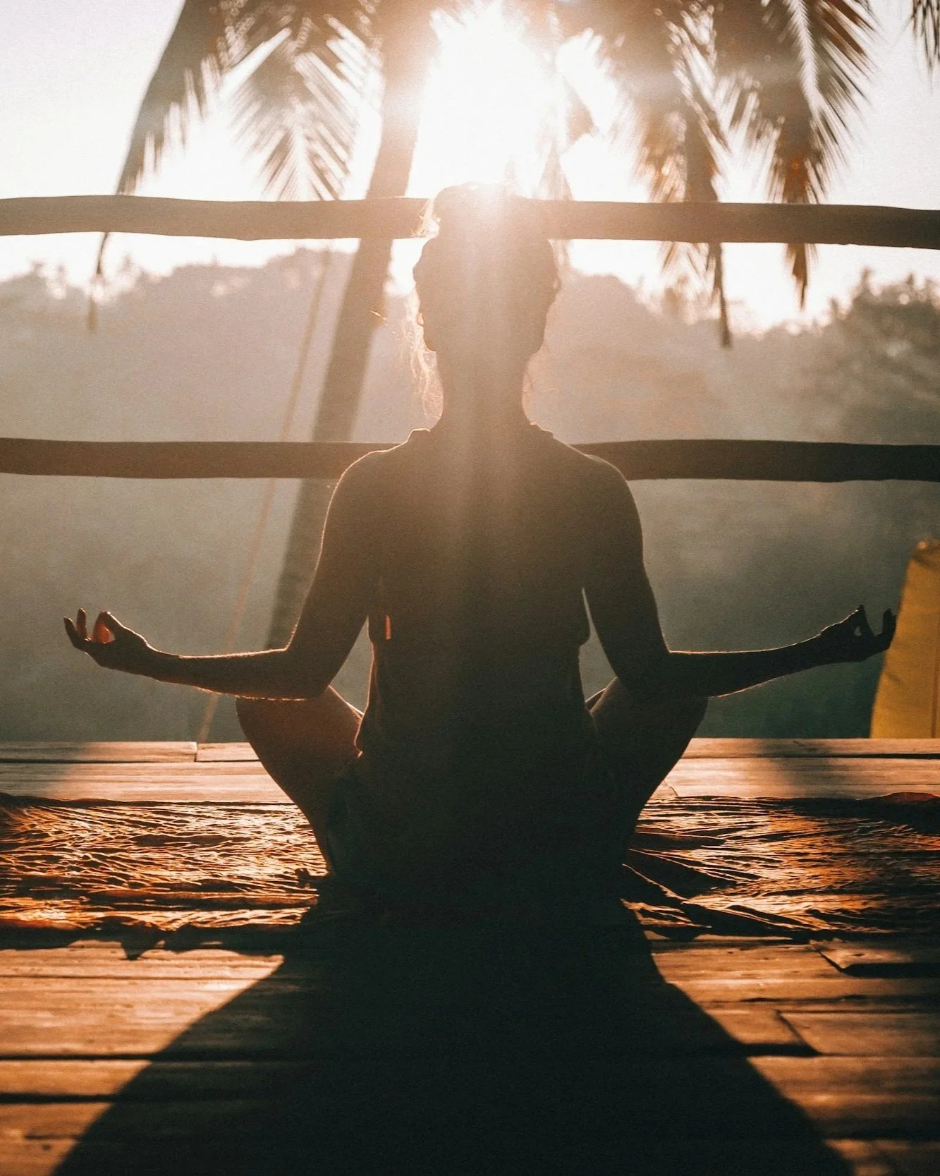 A person practicing yoga outdoors during sunset, sitting on a wooden deck with a tropical backdrop of palm trees, in a lotus position with hands resting on knees.