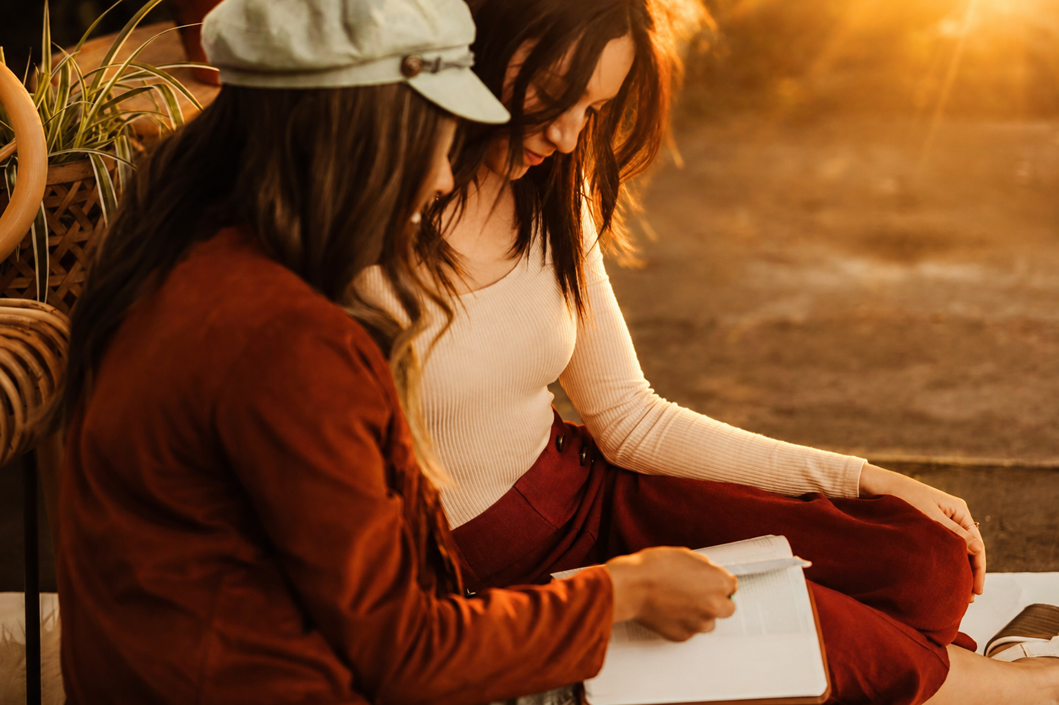 Two women sitting outdoors at sunset, looking at a book together, one wearing a beige sweater and the other in a brown jacket and gray cap, near potted plants.