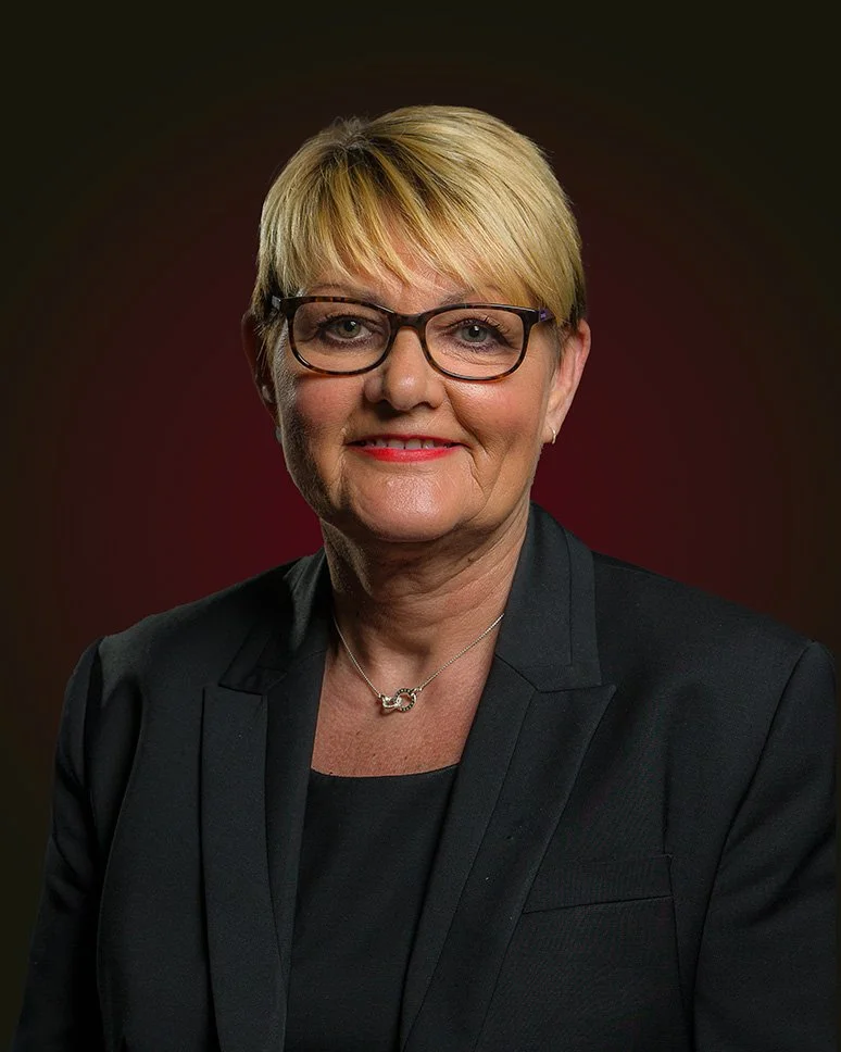 Portrait of a woman with short blonde hair, wearing glasses and a black suit, set against a dark backdrop.