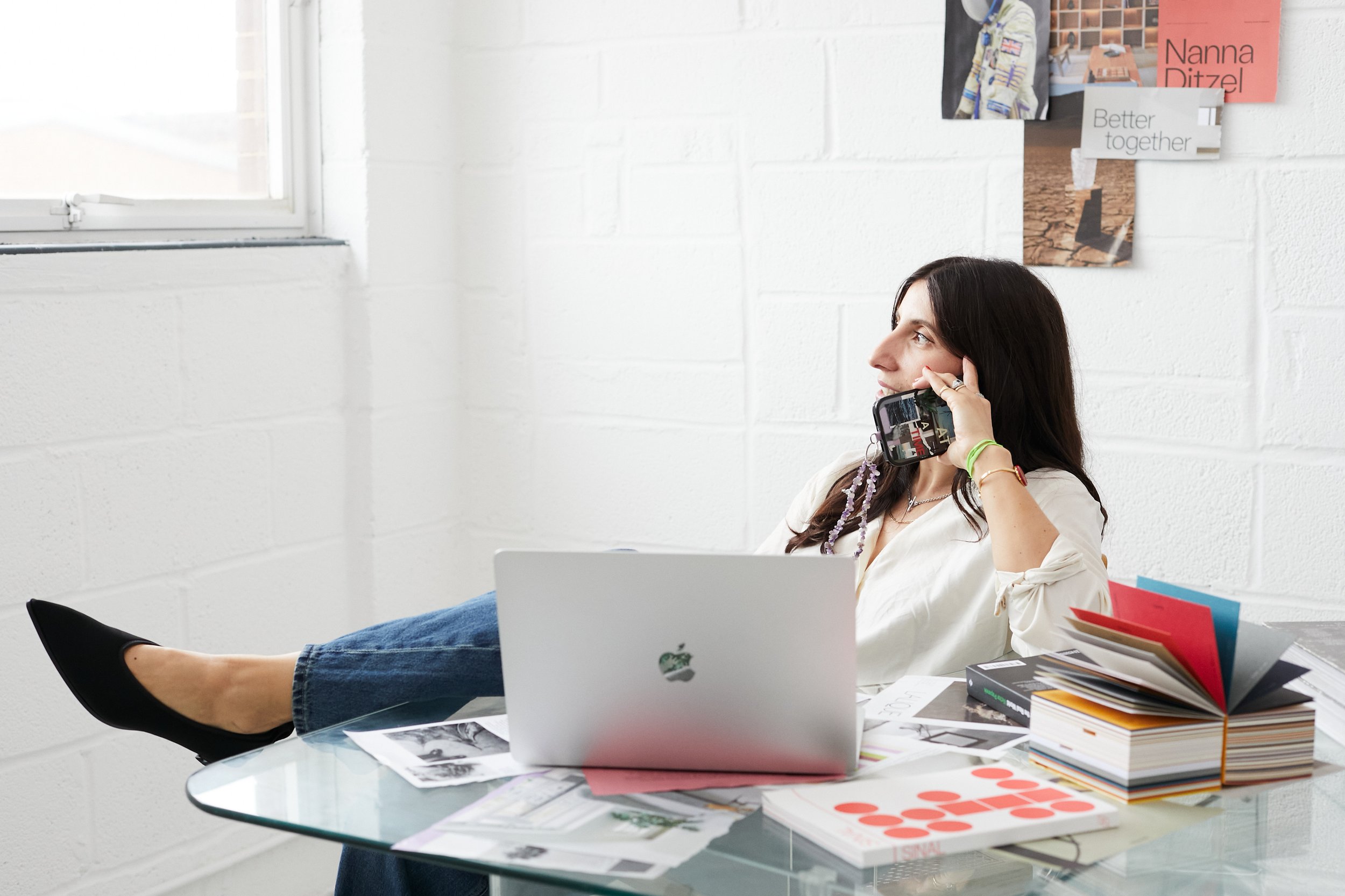 Creative Director Woman sitting in her studio at a cluttered desk with a laptop, talking on a phone, with an open book and folders on the desk, in a bright room with white brick wall and window. Ferrgood Studio.