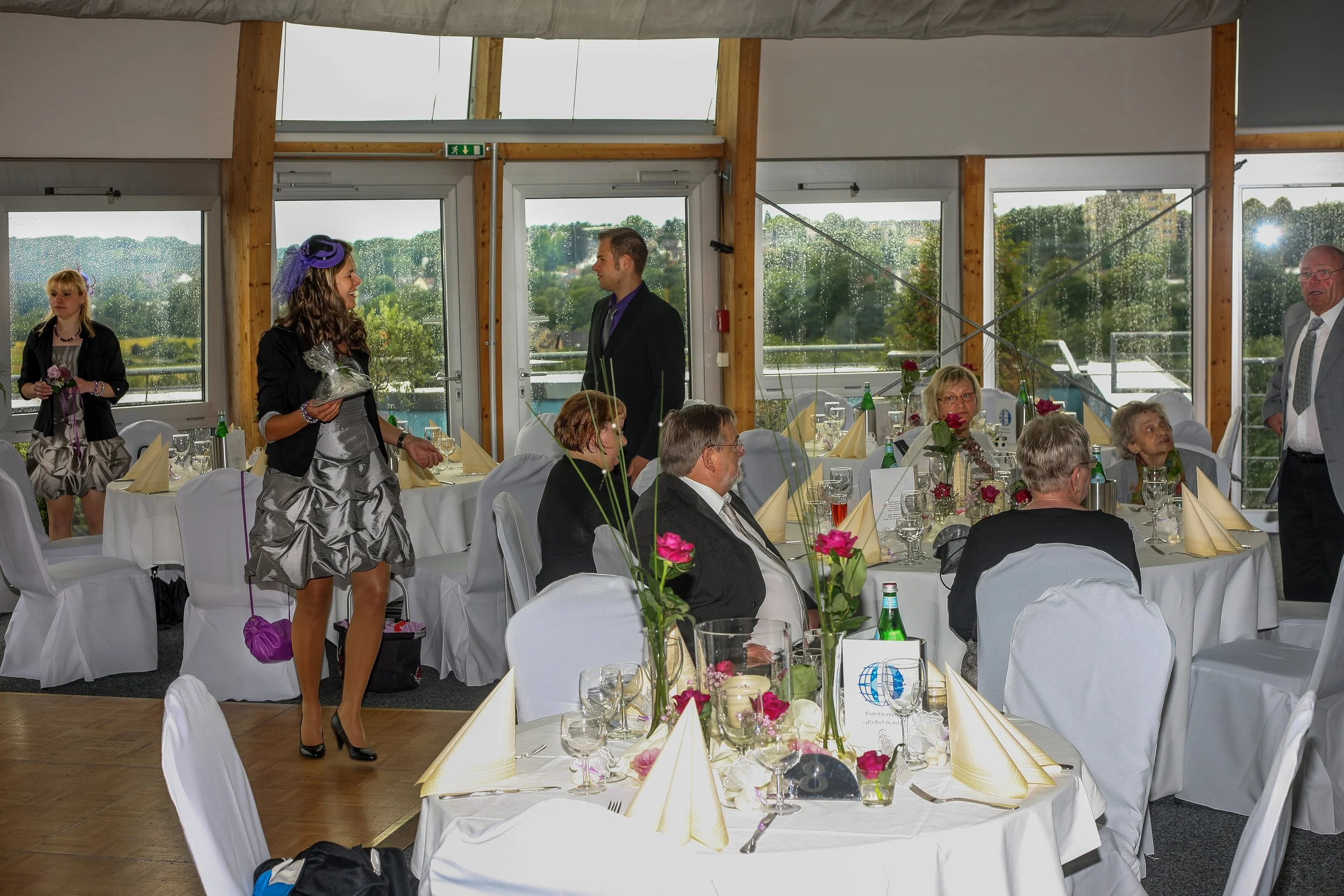 Menschen bei einem festlichen Anlass, einige sitzen am Tisch, andere stehen und unterhalten sich. Es handelt sich um eine festliche Veranstaltung in einem hellem Raum mit großen Fenstern, durch die man eine grüne Landschaft sieht.