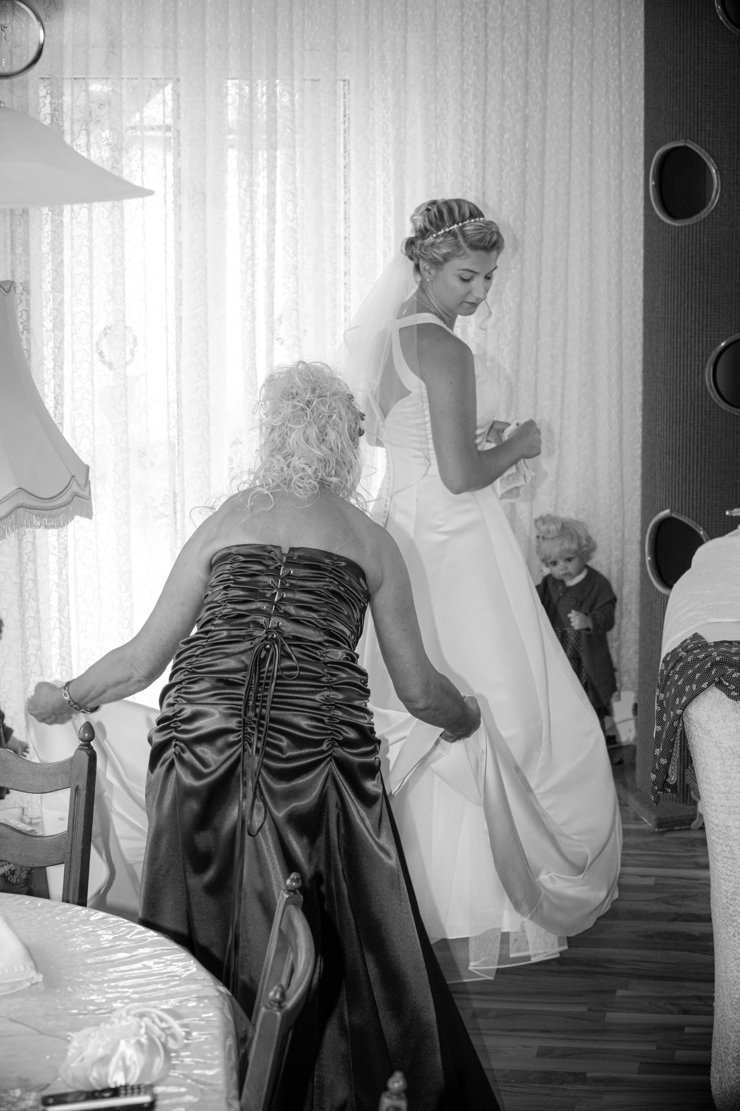 Eine Braut in einem weißen Hochzeitkleid mit einem Schleier steht im Hintergrund, während eine Frau in einem dunklen Kleid Gegenstände auf den Boden legt. Es gibt Kinder und andere erwachsene Gäste im Raum, die bei der Hochzeit beteiligt sind.