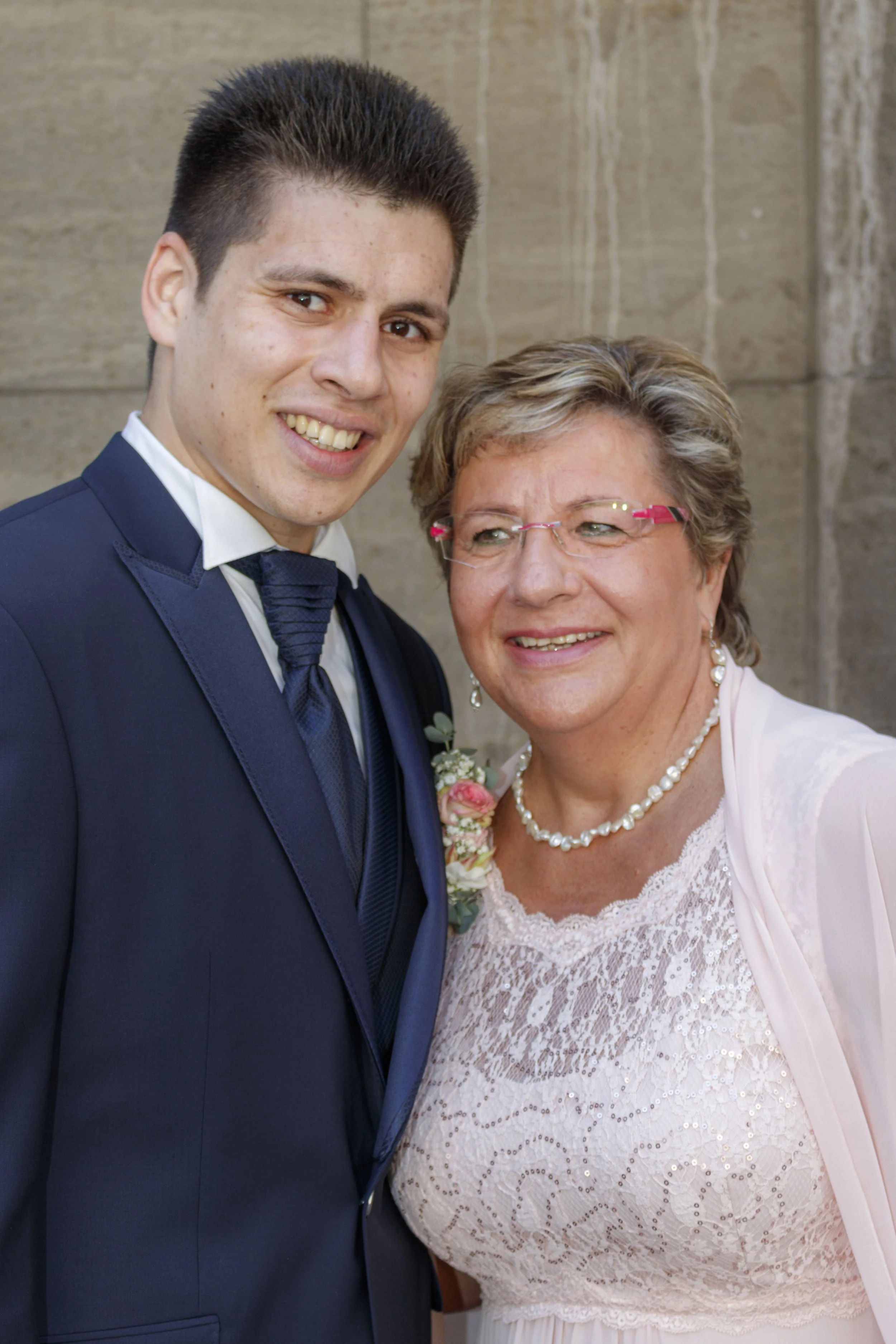 Junge und ältere Frau bei einer Feier, vermutlich Hochzeit, lächeln sich freundlich an, an einem Steingebäude.