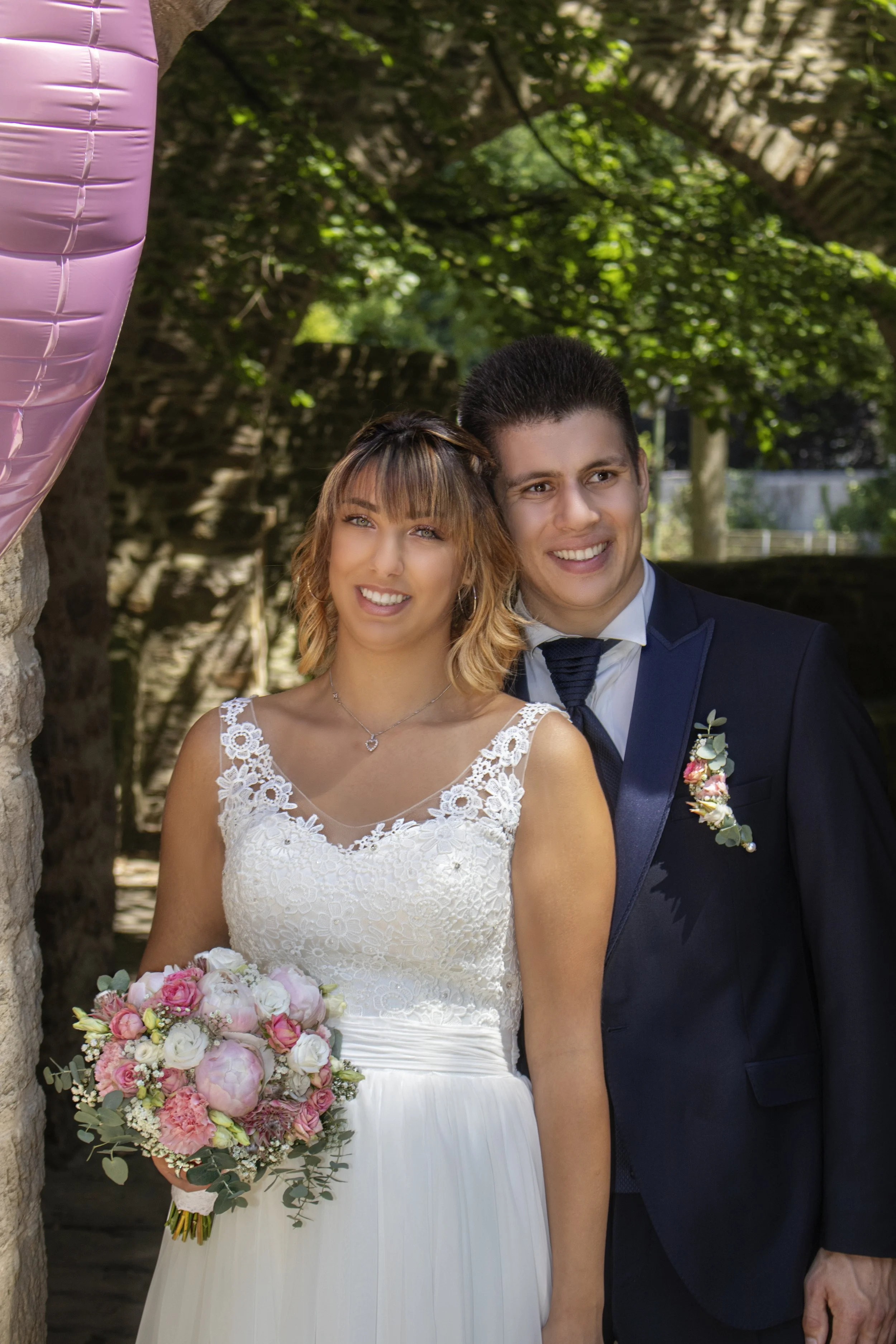 Ein Brautpaar steht nebeneinander im Freien bei Tageslicht. Die Braut trägt ein weißes Hochzeitskleid mit Spitzenmuster und hält einen großen Blumenstrauß aus pinken und weißen Blumen. Der Bräutigam trägt einen dunklen Anzug mit Lackschuhen. Hinter i