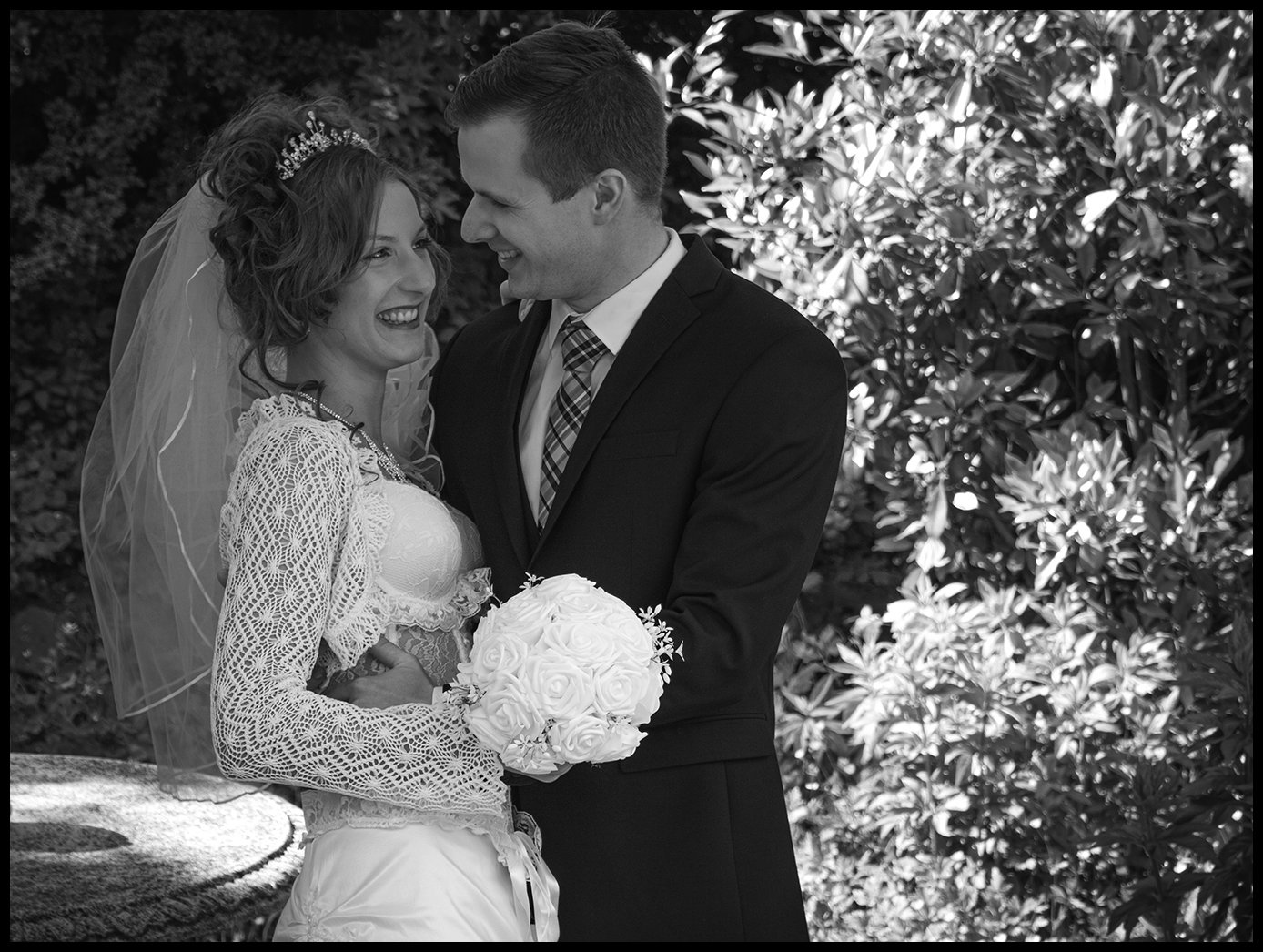 Braut und Bräutigam freuen sich bei ihrer Hochzeit im Freien, die Braut hält einen Blumenstrauß, beide lachen, im Hintergrund grünen Blätter.
