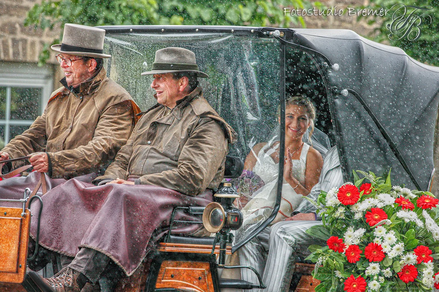 Eine Hochzeitskutsche mit vier Menschen, die bei Regen fahren. Die Fahrer sind in brauner Kleidung und Hüten, die Braut sitzt hinten und winkt. Es gibt einen großen Blumenstrauß mit roten und weißen Blumen. Der Hintergrund zeigt grüne Bäume und eine 