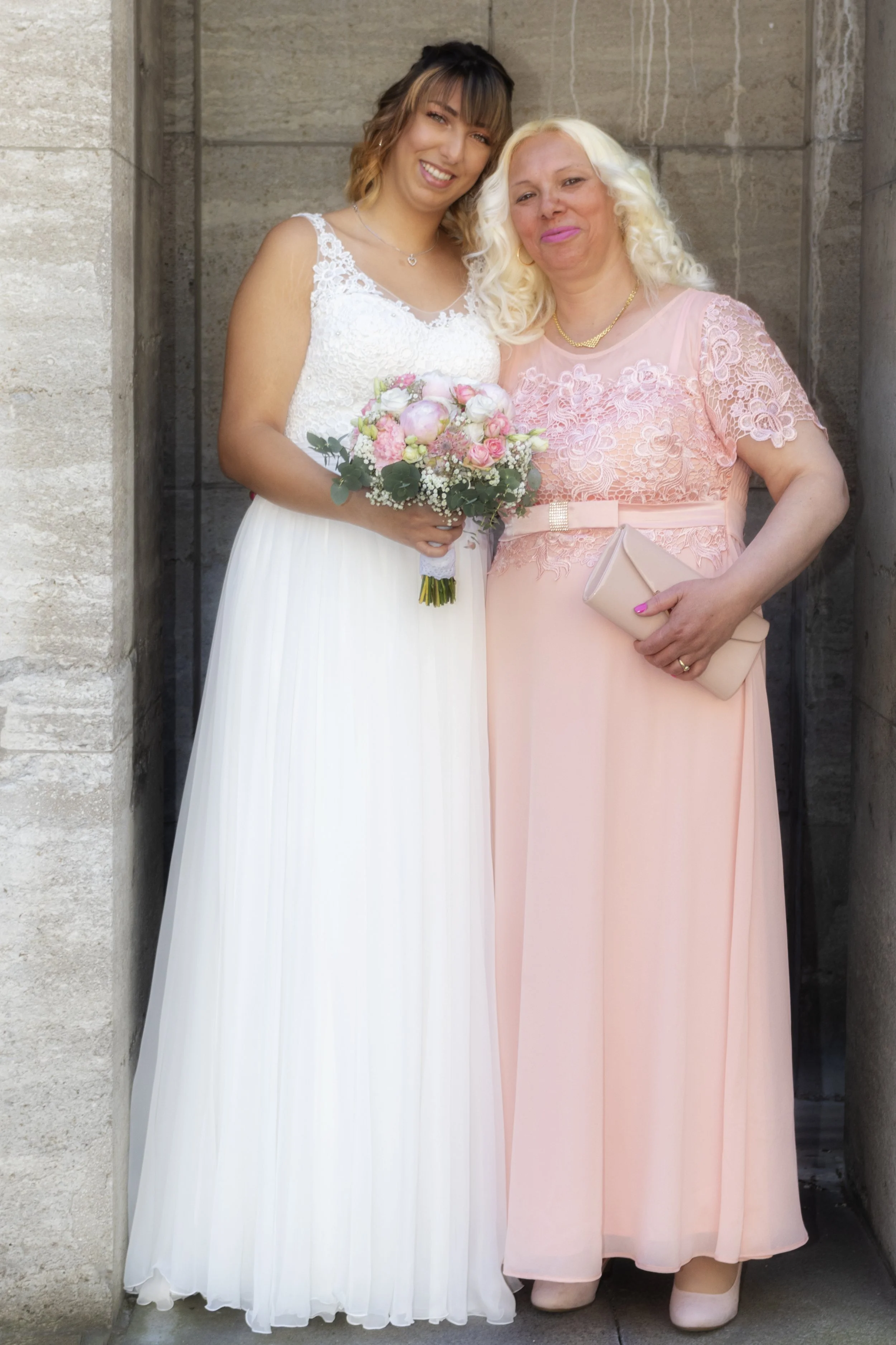 Zwei Frauen stehen nebeneinander vor einer Steinfassade, eine trägt ein weißes Brautkleid mit Blumenstrauß, die andere ein hellrosa Kleid mit Clutch und Goldkette.