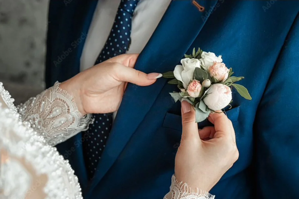 Nahaufnahme einer Frau, die einem Mann ein Blumenröckchen an die Brust steckt. Der Mann trägt einen blauen Anzug und ein weißes Hemd, die Frau trägt eine Spitze mit Perlen. Es handelt sich wahrscheinlich um eine Hochzeitszeremonie.