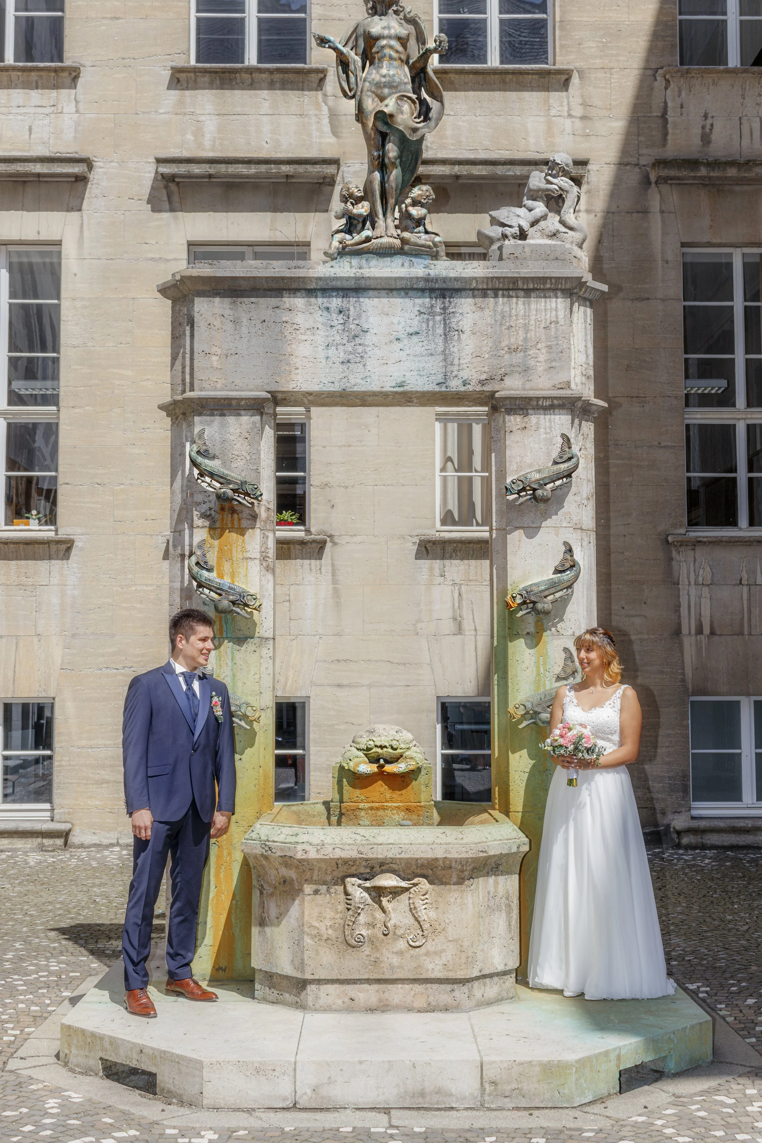 Ein Brautpaar steht vor einem historischen Brunnen mit Wasserspeiern in einer Stadt. Der Bräutigam trägt einen dunklen Anzug, die Braut hält einen Blumenstrauß. Es ist ein sonniger Tag.
