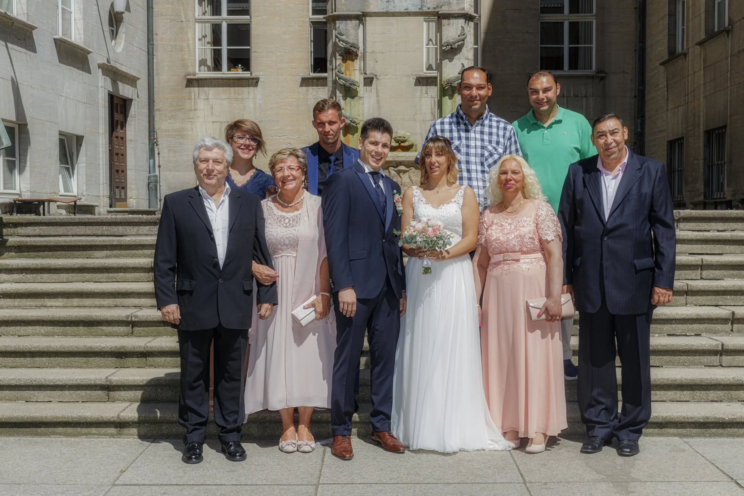 Eine Hochzeitsgruppe, bestehend aus mehreren Männern und Frauen, steht auf Stufen vor einem historischen Gebäude mit Steinfassade und Wasserspeiern. Das Brautpaar befindet sich in der Mitte, sie trägt ein weißes Hochzeitskleid und hält einen Blumenst