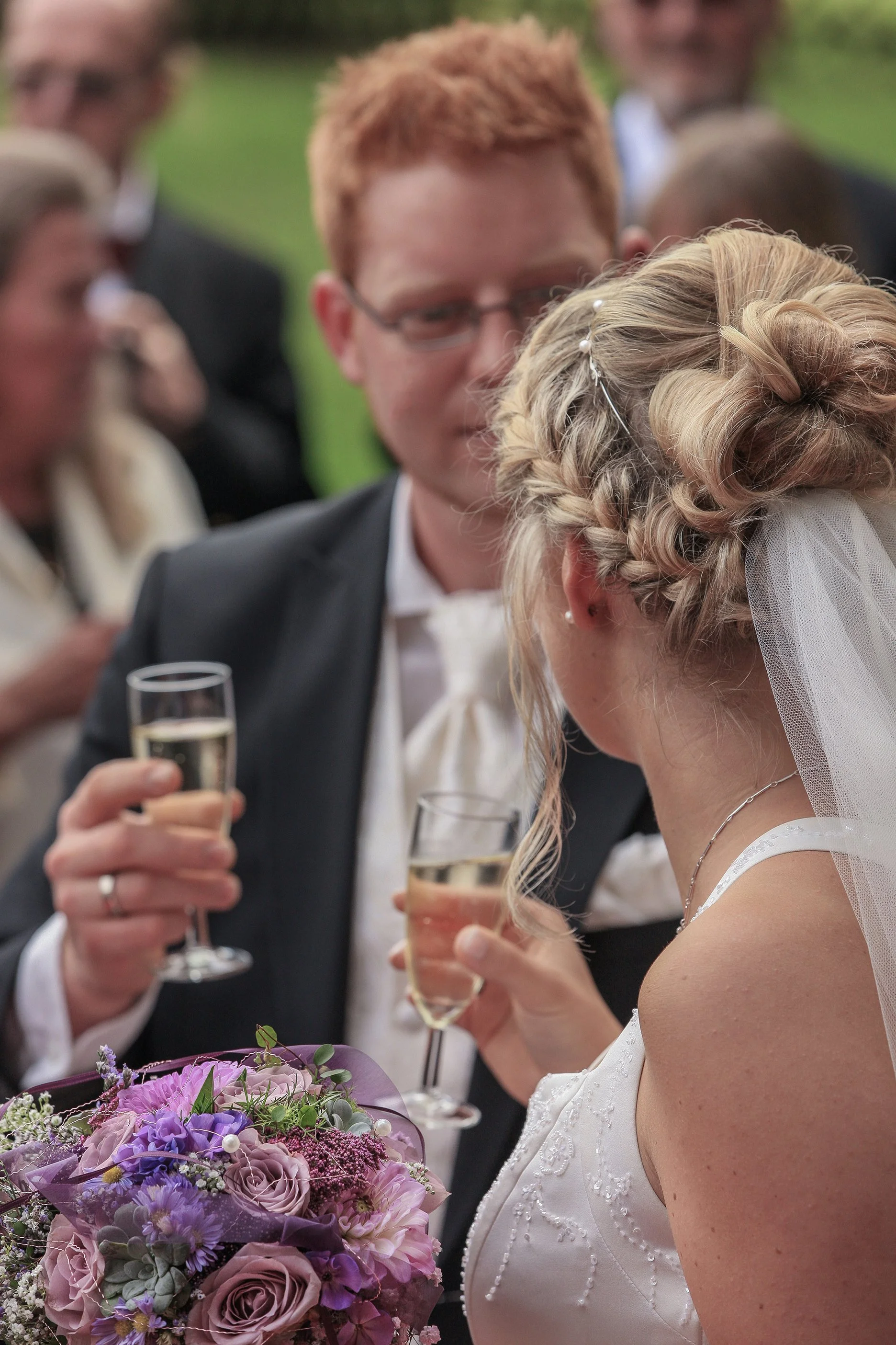 Ein Brautpaar beim Trinken mit Freunden bei einer Hochzeit, mit Blumenstrauß im Vordergrund.