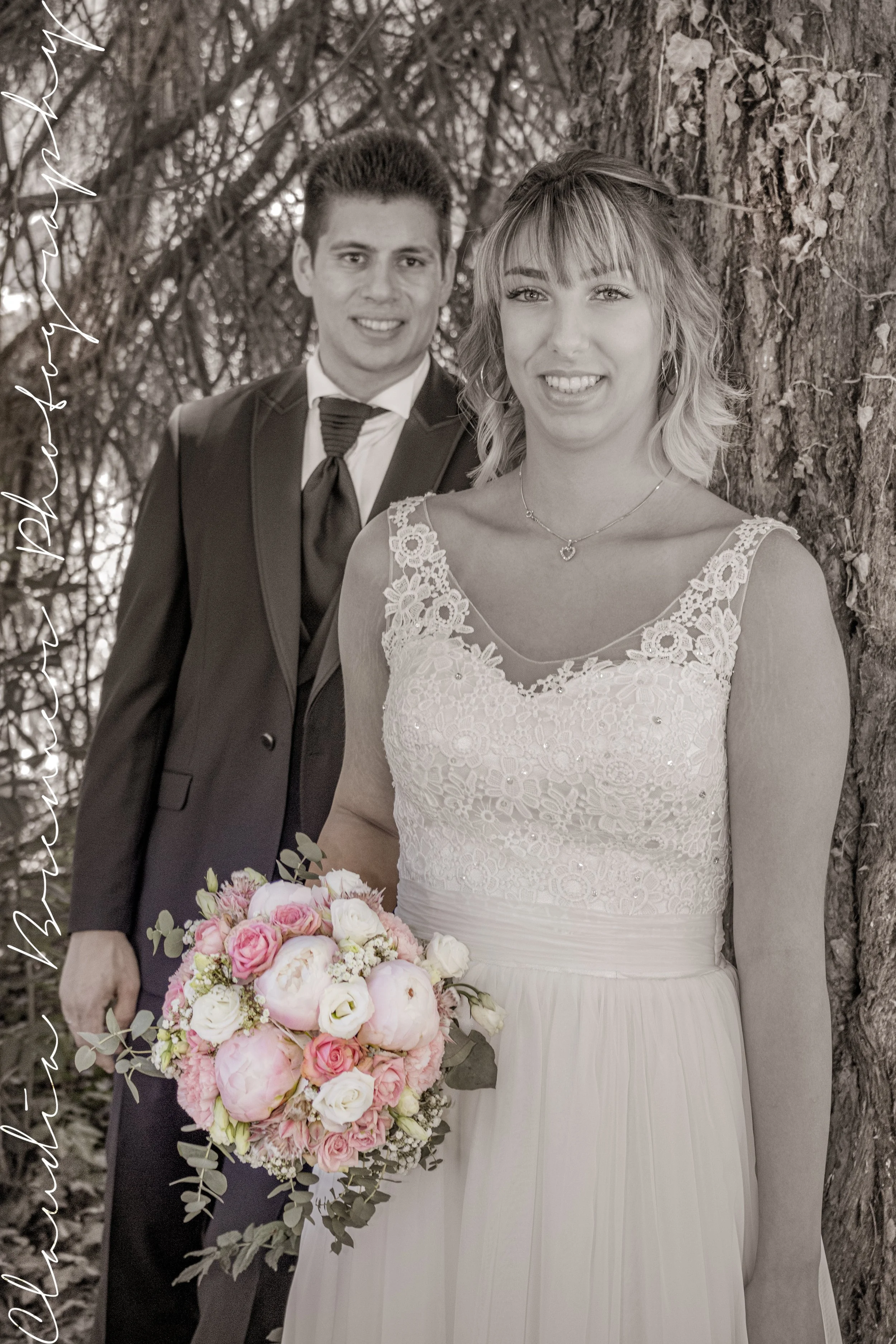Ein Brautpaar steht zusammen im Freien vor einem Baum, die Braut trägt ein weises Hochzeitskleid mit Lace-Details und hält einen großen Blumenstrauß, der Bräutigam trägt eine dunkle Anzug mit Krawatte.