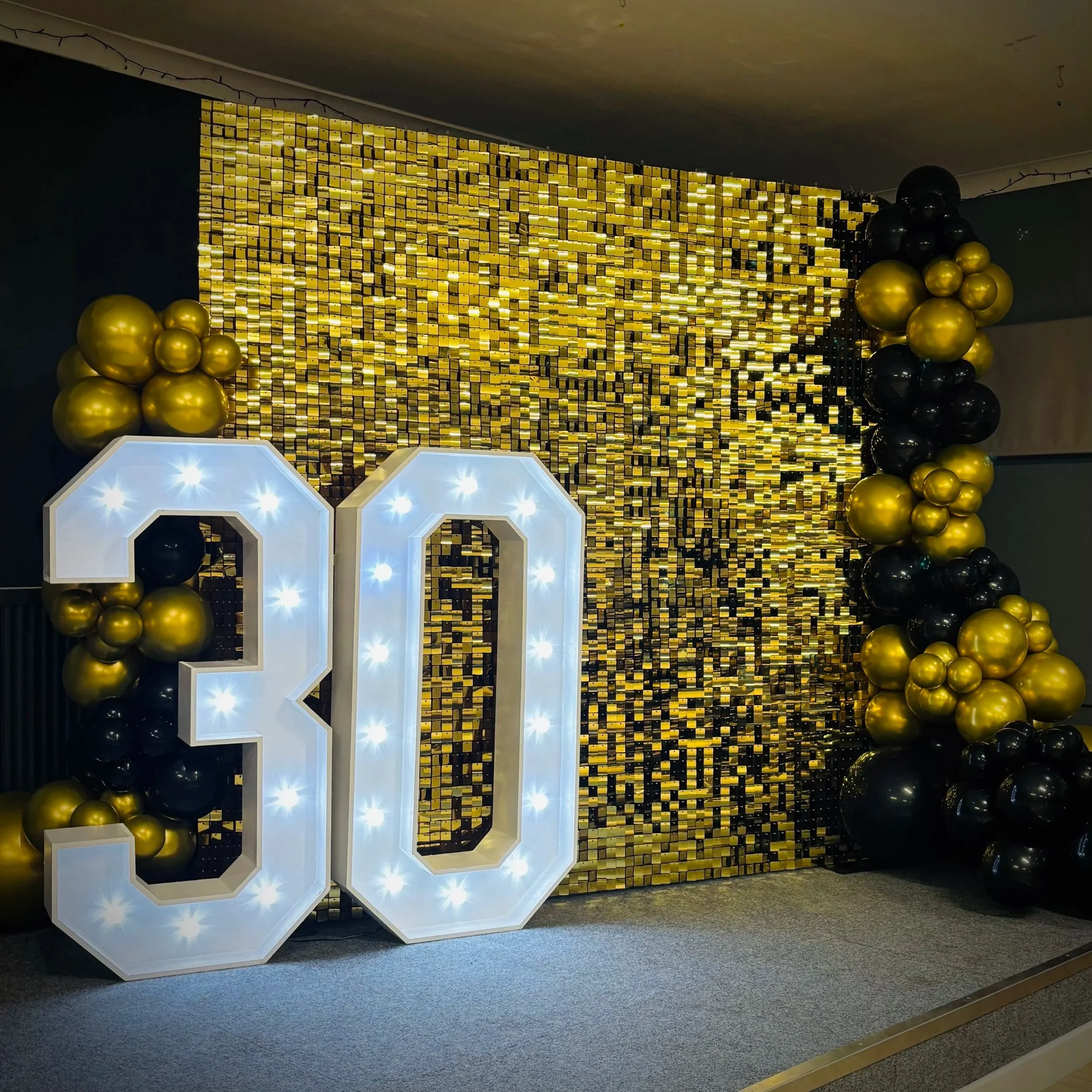 Large light up number display reading '2023' positioned on a light wooden floor, with white balloon decorations on either side, against a white brick wall backdrop, likely for a New Year's celebration or event.