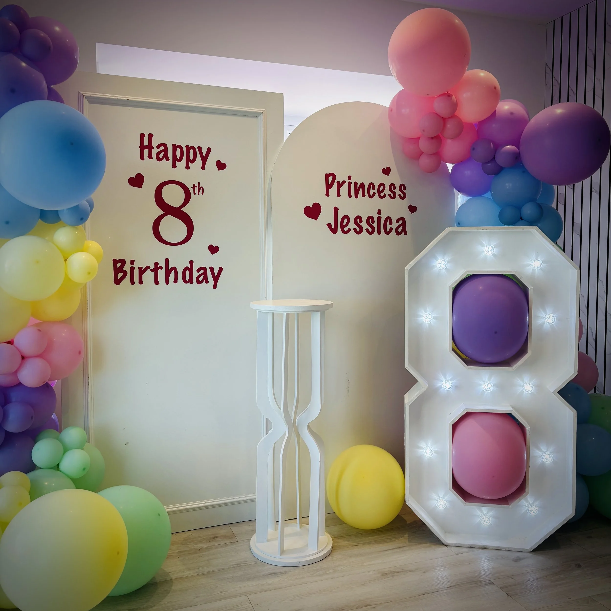 A white backdrop with red cursive text that says 'I was always you', decorated with a cluster of white balloons on the left side, in a room with wooden furniture, curtains, and large windows showing a golf cart and trees outside.