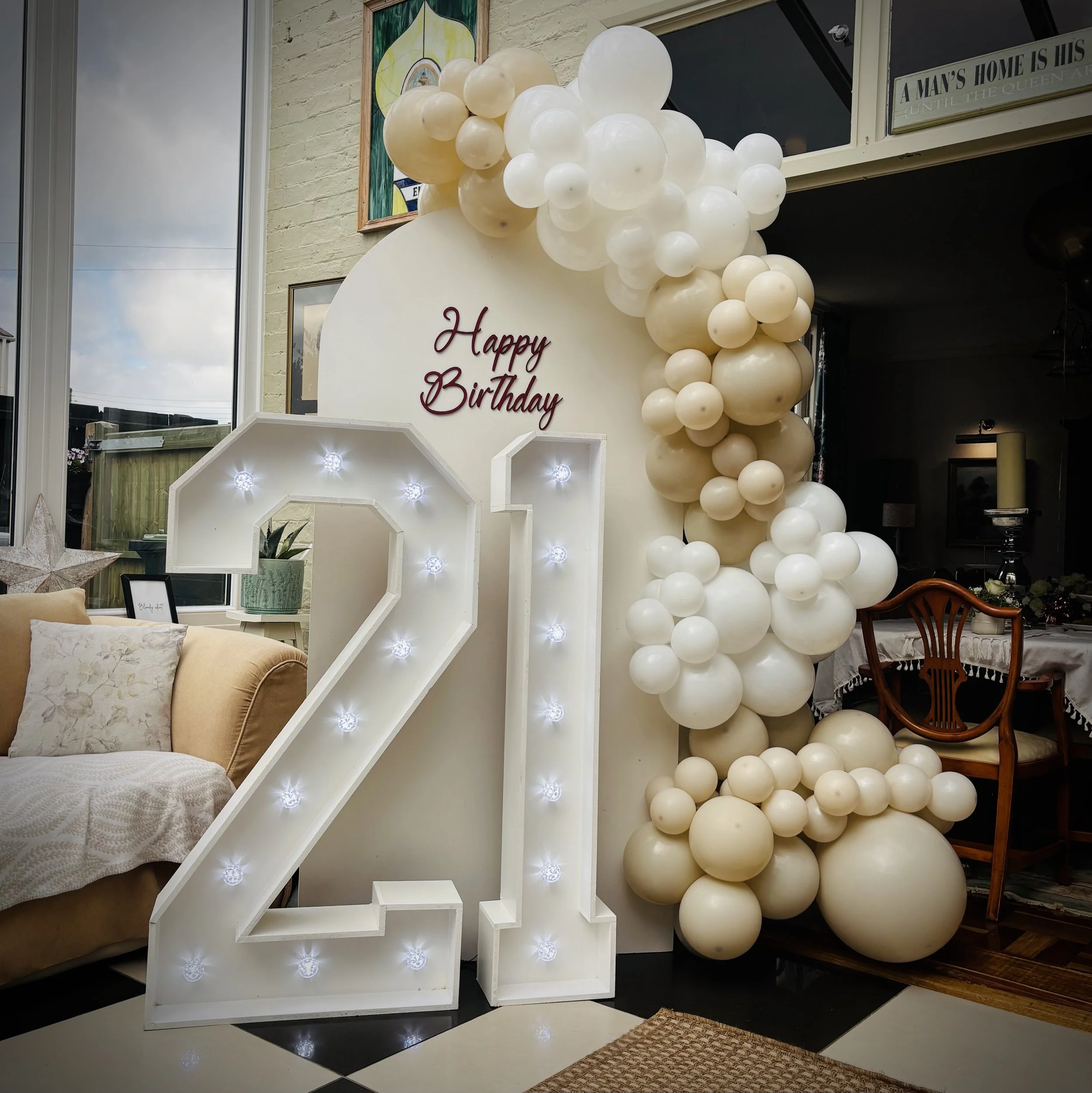 Decorated birthday celebration display with a large illuminated number 21, a white backdrop with the words "Happy Birthday" in red, and an arch of balloons in shades of white and cream. There is a sofa with pillows and a side table with plants, along with a dining area in the background.