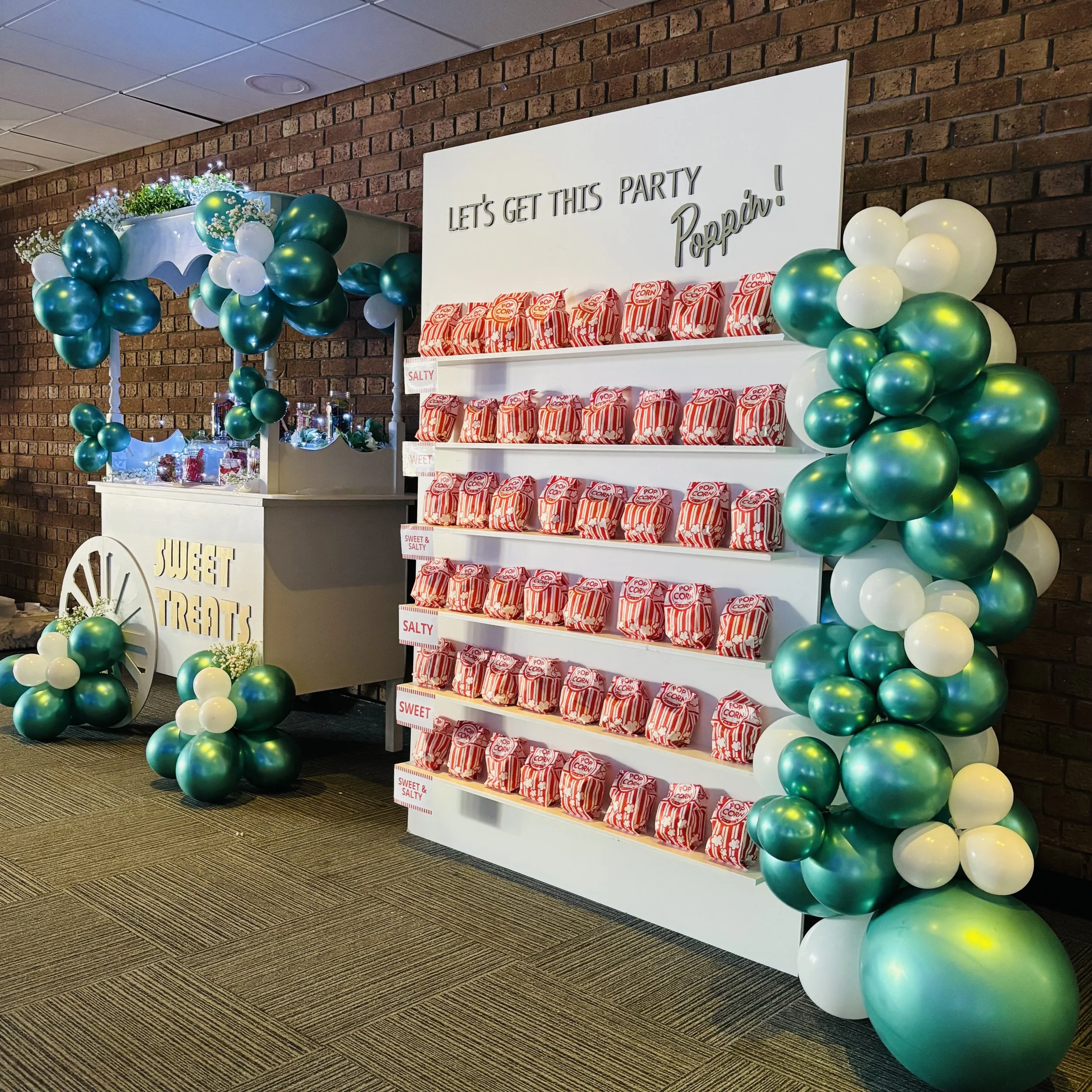 Decorative party display with a white cart labeled "Sweet Treats" decorated with teal, white, and gold balloons, next to a popcorn stand with multiple boxes of popcorn on shelves, against a brick wall and a brown patterned carpet.