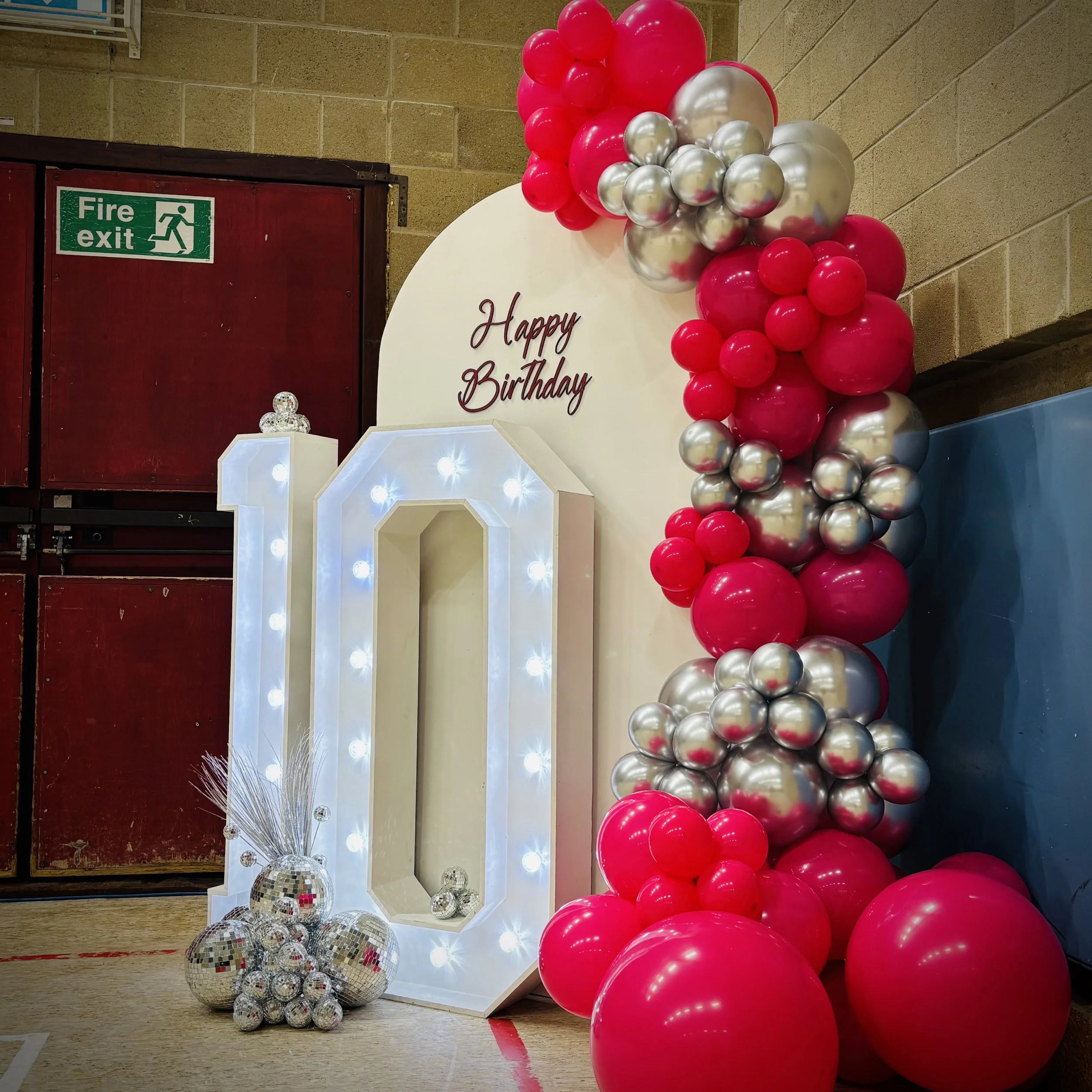A white backdrop with red cursive text that says 'I was always you', decorated with a cluster of white balloons on the left side, in a room with wooden furniture, curtains, and large windows showing a golf cart and trees outside.