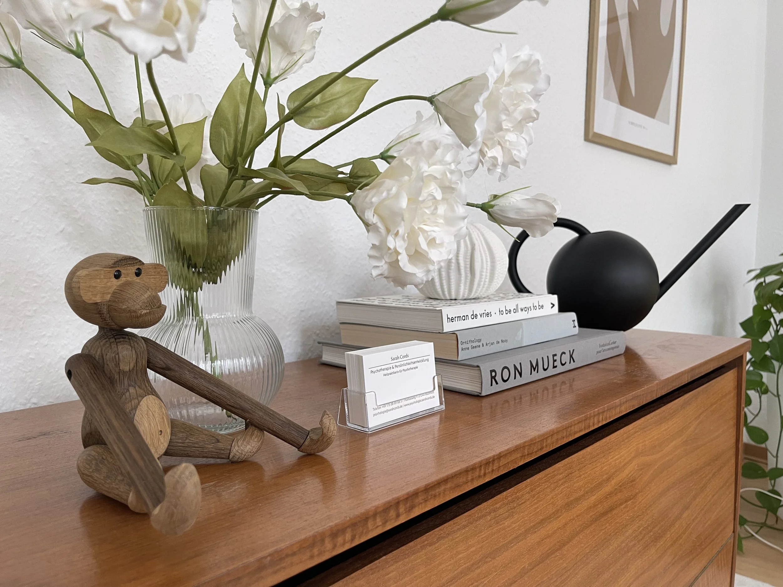 Auf einer Kommode steht ein Strauß weißer Blumen, ein Buchstapel, eine Gießkanne und ein Kai Bojesen Affe aus Holz