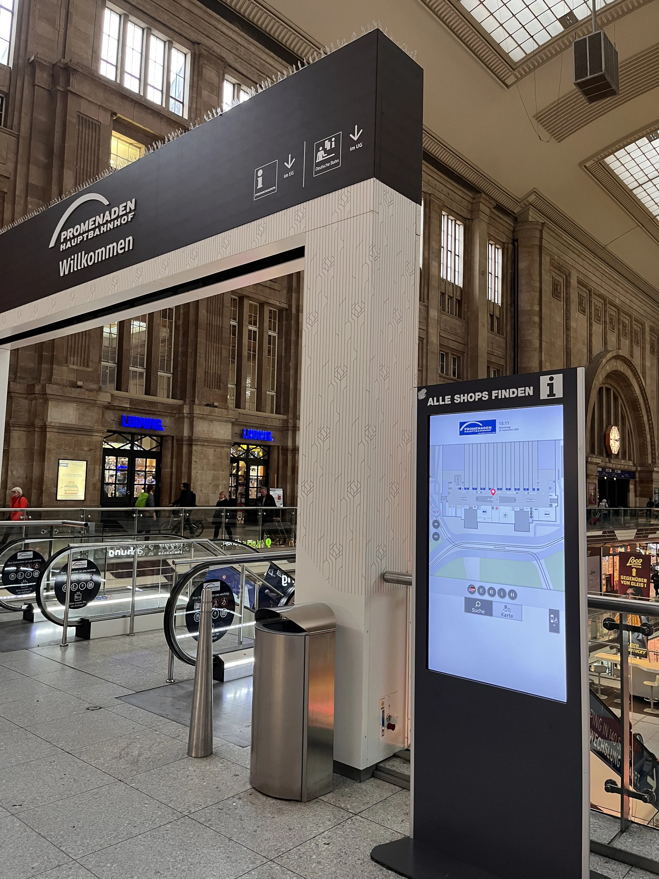 Innenansicht eines Bahnhofs mit Aufzug, Informationsstand und digitaler Karte, Beschilderung für Geschäfte und Hinweise auf den Bahnhof in Deutschland.