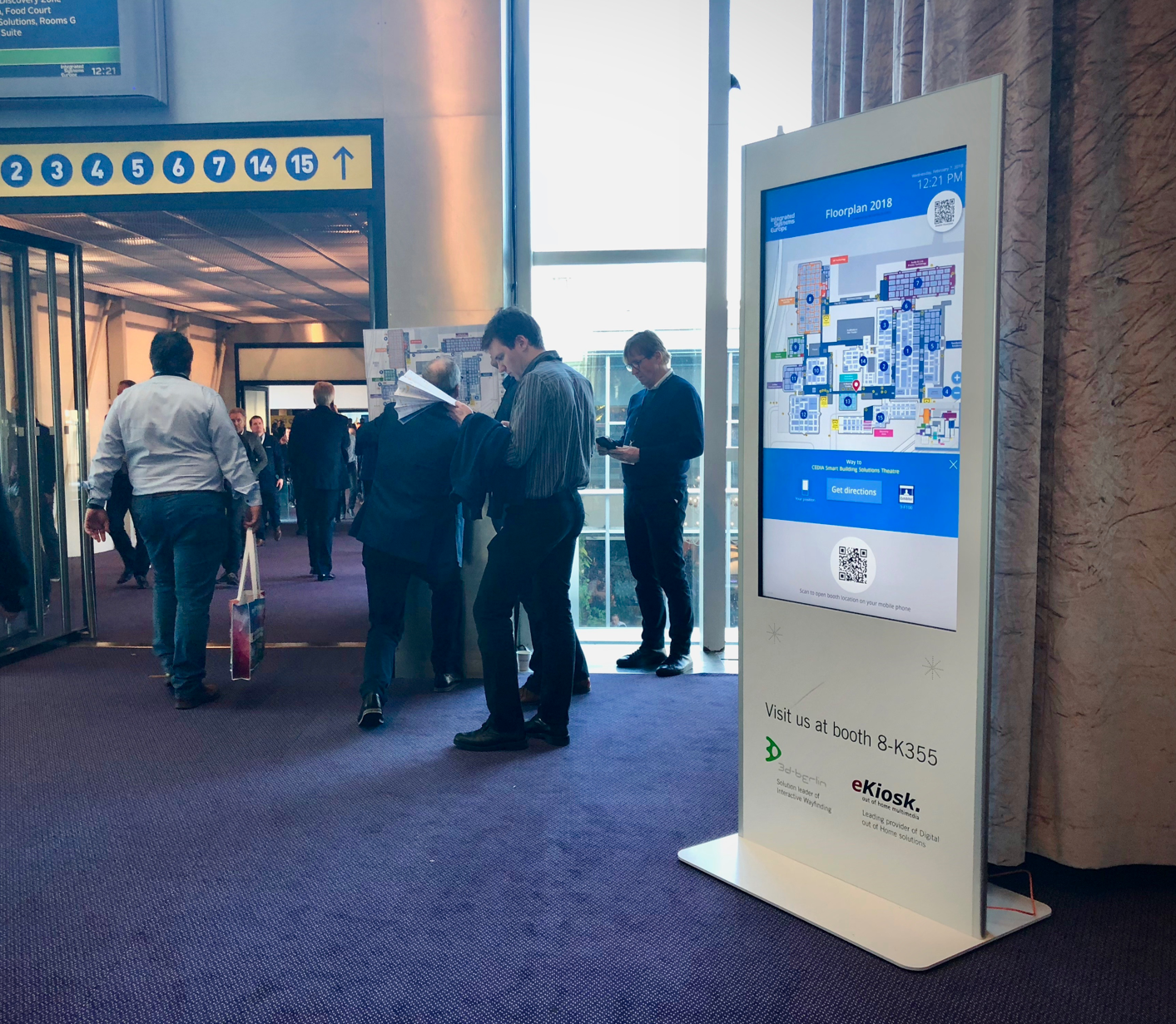 Menschen stehen in einem Flur bei einer digitalen Wegbeschreibungstafel in einer Konferenzhalle.