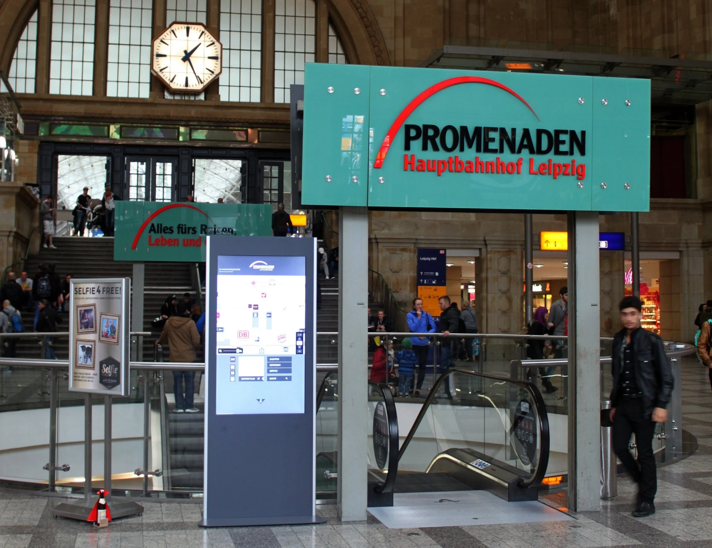 Innenaufnahme des Leipziger Hauptbahnhofs mit einem großen Schild, das auf den Ausgang 'PROMENADEN Hauptbahnhof Leipzig' hinweist. Im Hintergrund sind Menschen, Treppen und Geschäfte sichtbar.