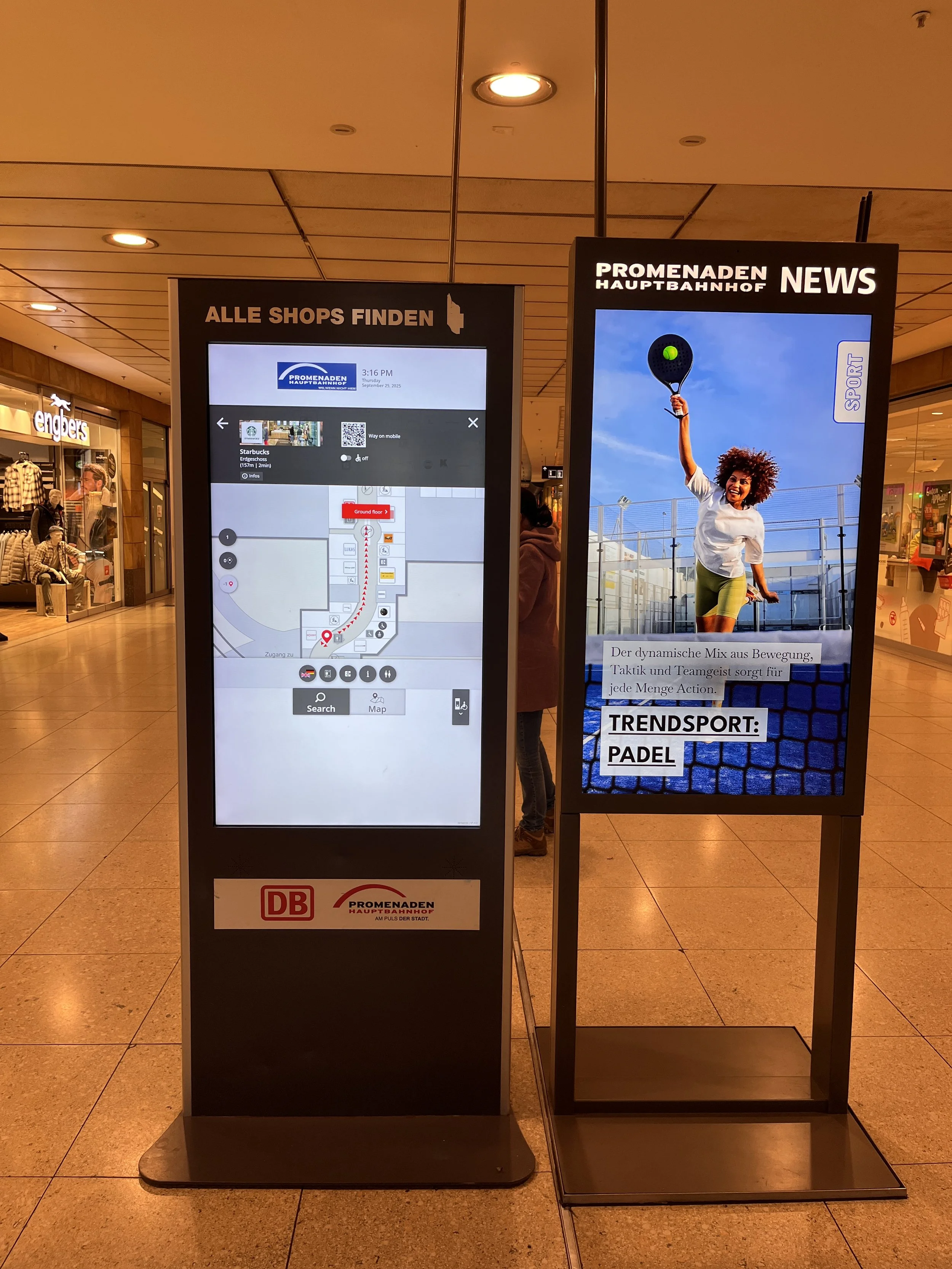 Zwei interaktive Anzeigetafeln in einem Einkaufszentrum. Die linke zeigt eine Karte des Einkaufszentrums mit Geschäften, die gefunden werden können, die rechte bewirbt Tennis und Padel als Trendsportarten mit einem Bild einer Frau, die Padel spielt.