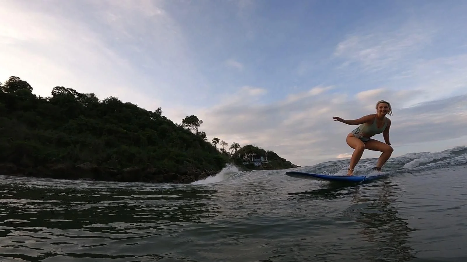 A woman surfing on a wave near a lush, green hillside during sunset.
